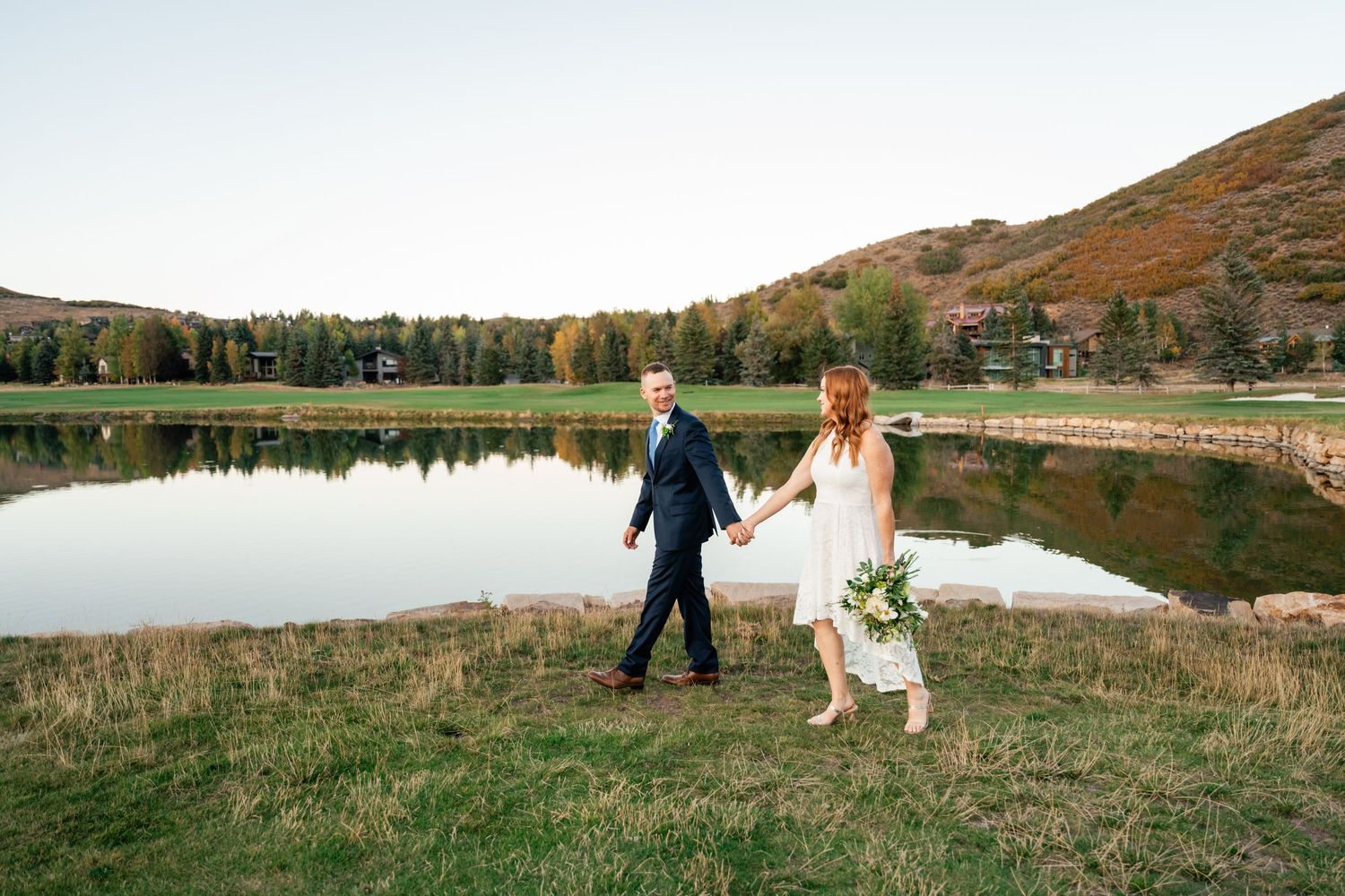 Two people in wedding attire walk hand in hand by a serene mountain lake during autumn.