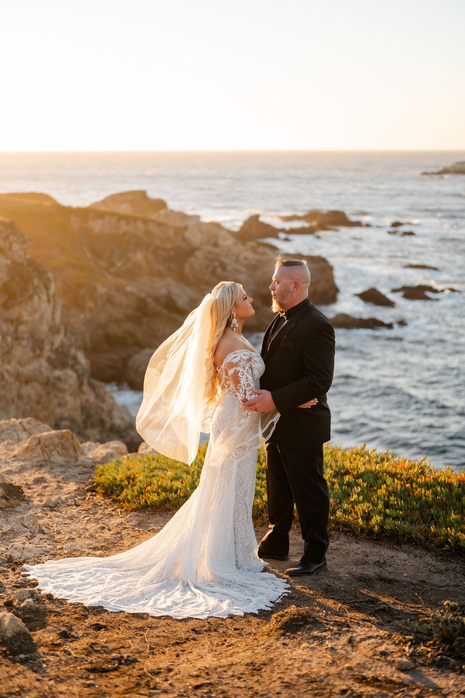 A couple poses on rocky cliffs overlooking the ocean during sunset.