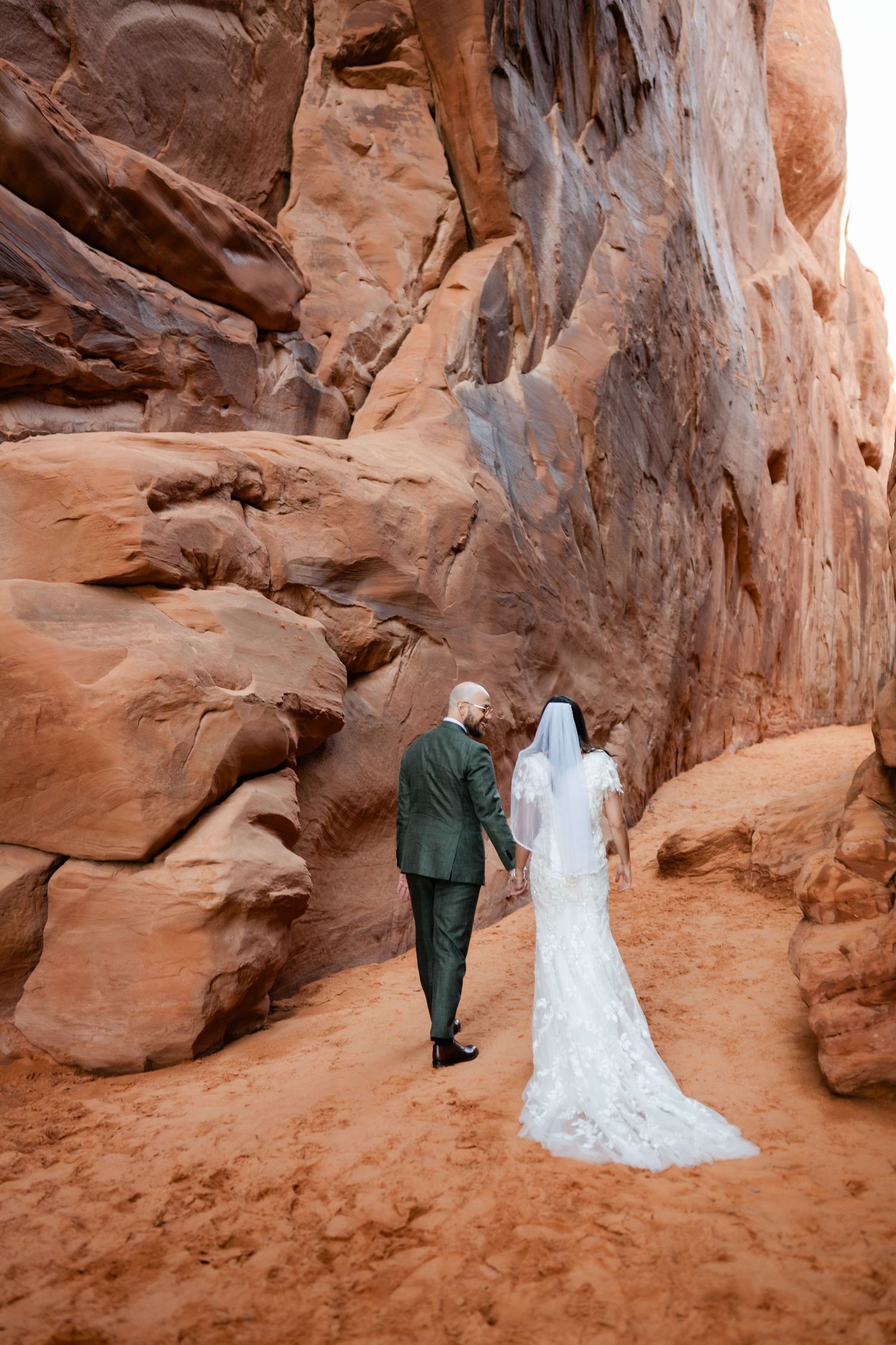 A couple in wedding attire walking through red rock canyon formations in the desert.