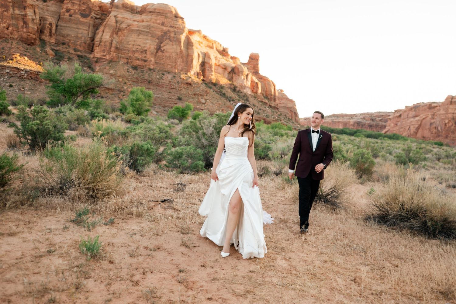 Bride and groom walk hand-in-hand through a scenic desert landscape with red rock formations at golden hour.