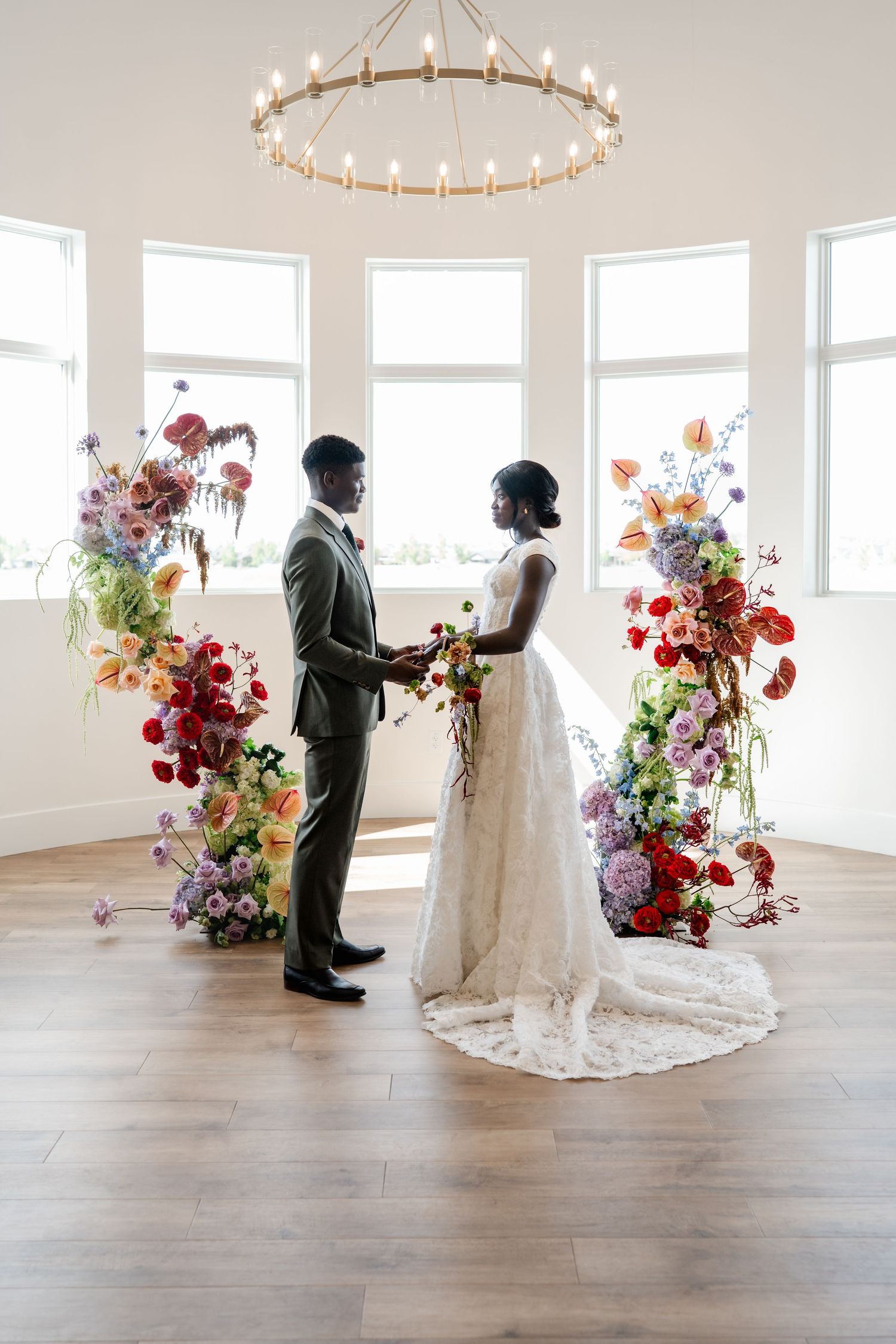 Bride and groom exchange vows at colorful floral arch ceremony in bright white venue with hardwood floors.