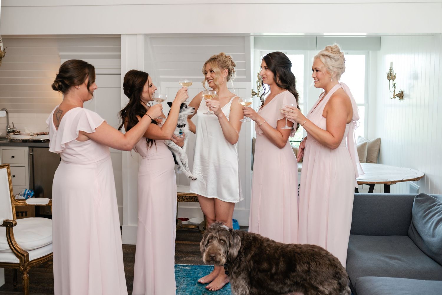 Bridesmaids in blush pink gowns toasting with the bride in white during wedding preparations, with a dog nearby.