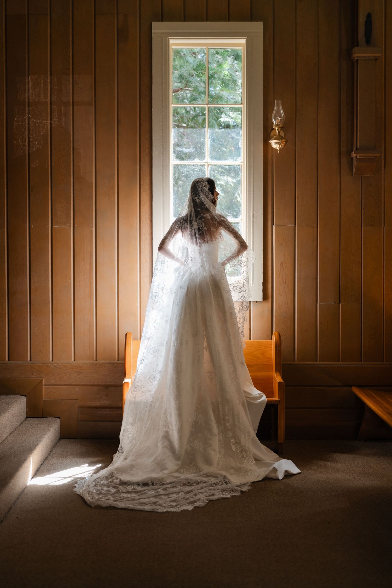 Bride in white lace wedding gown and veil stands before a sunlit window in a wooden chapel.