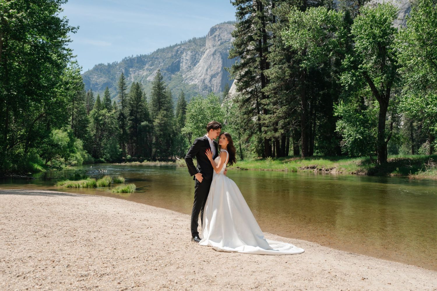 Bride and groom share a romantic dip kiss on a sandy riverbank surrounded by tall pine trees and mountains in Yosemite.