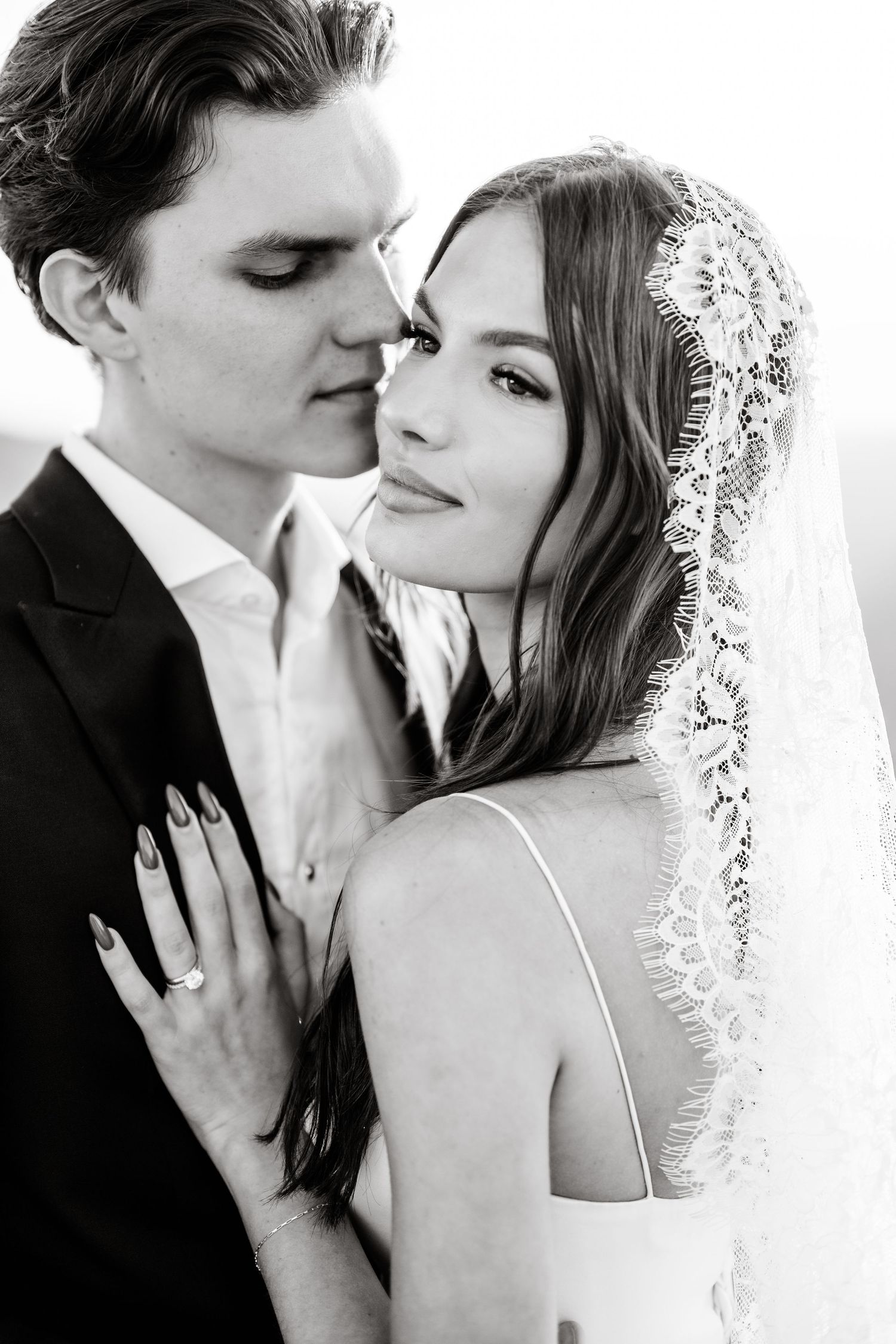 Black and white portrait of bride in lace veil and groom sharing intimate moment outdoors.