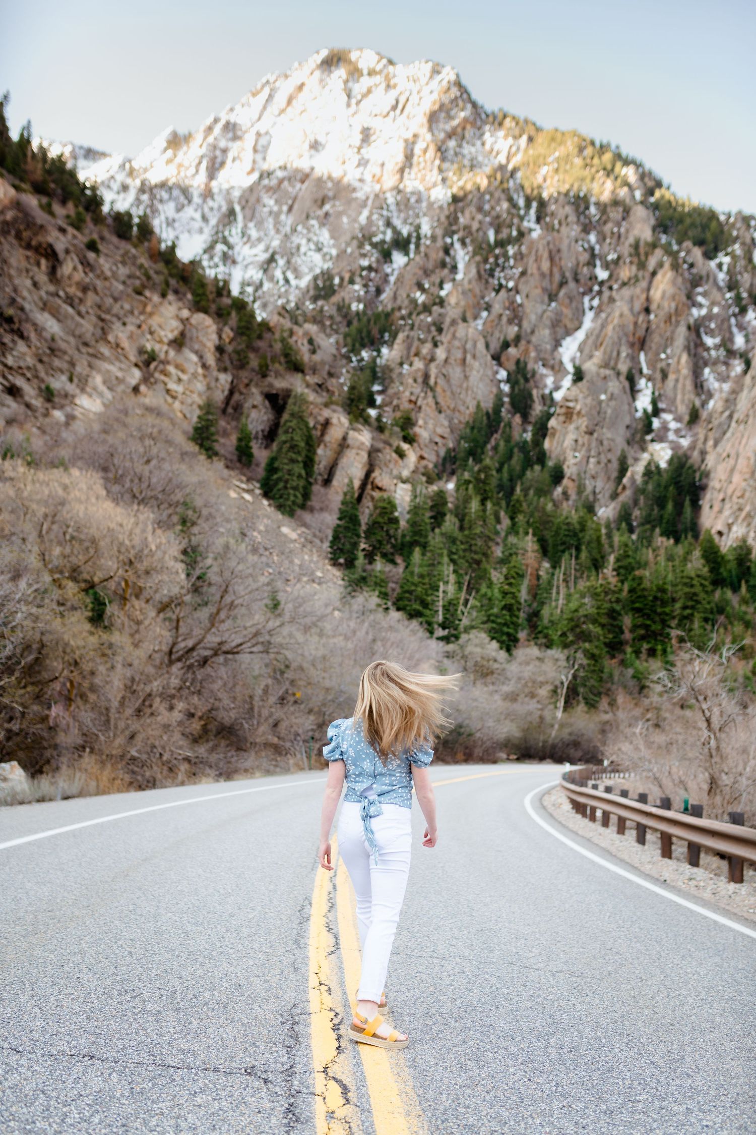 Person in light colored clothing walks along a winding mountain road with snow-capped peaks and evergreen trees in background.