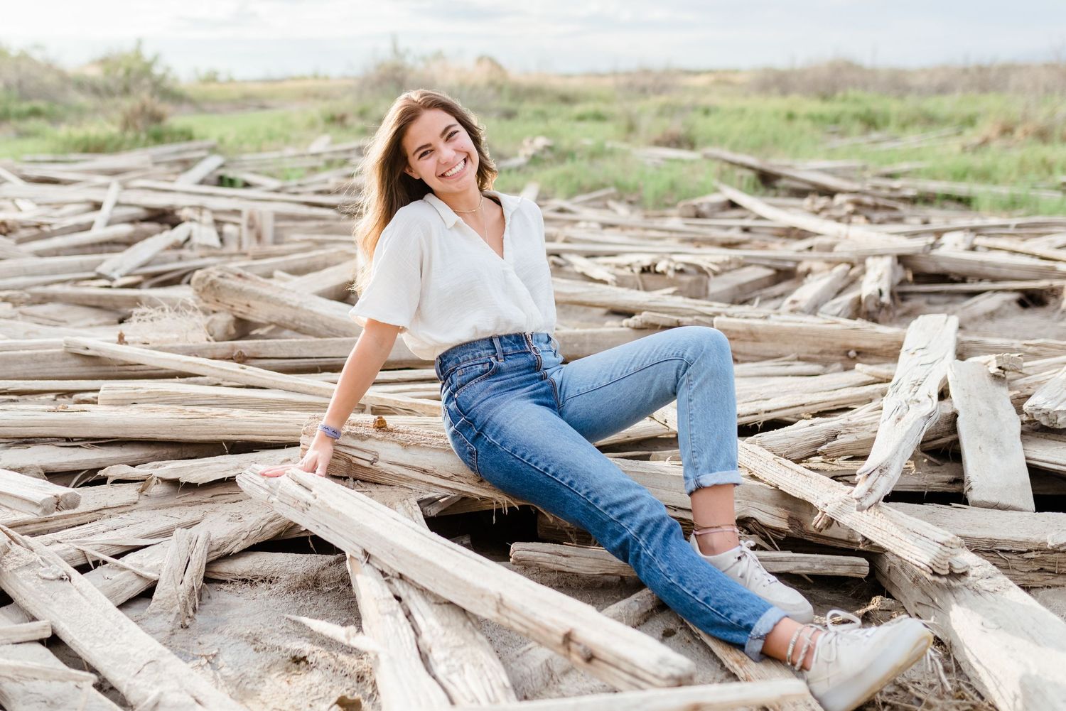 A person in blue jeans and white top reclines casually on driftwood at a beach during a sunny day.