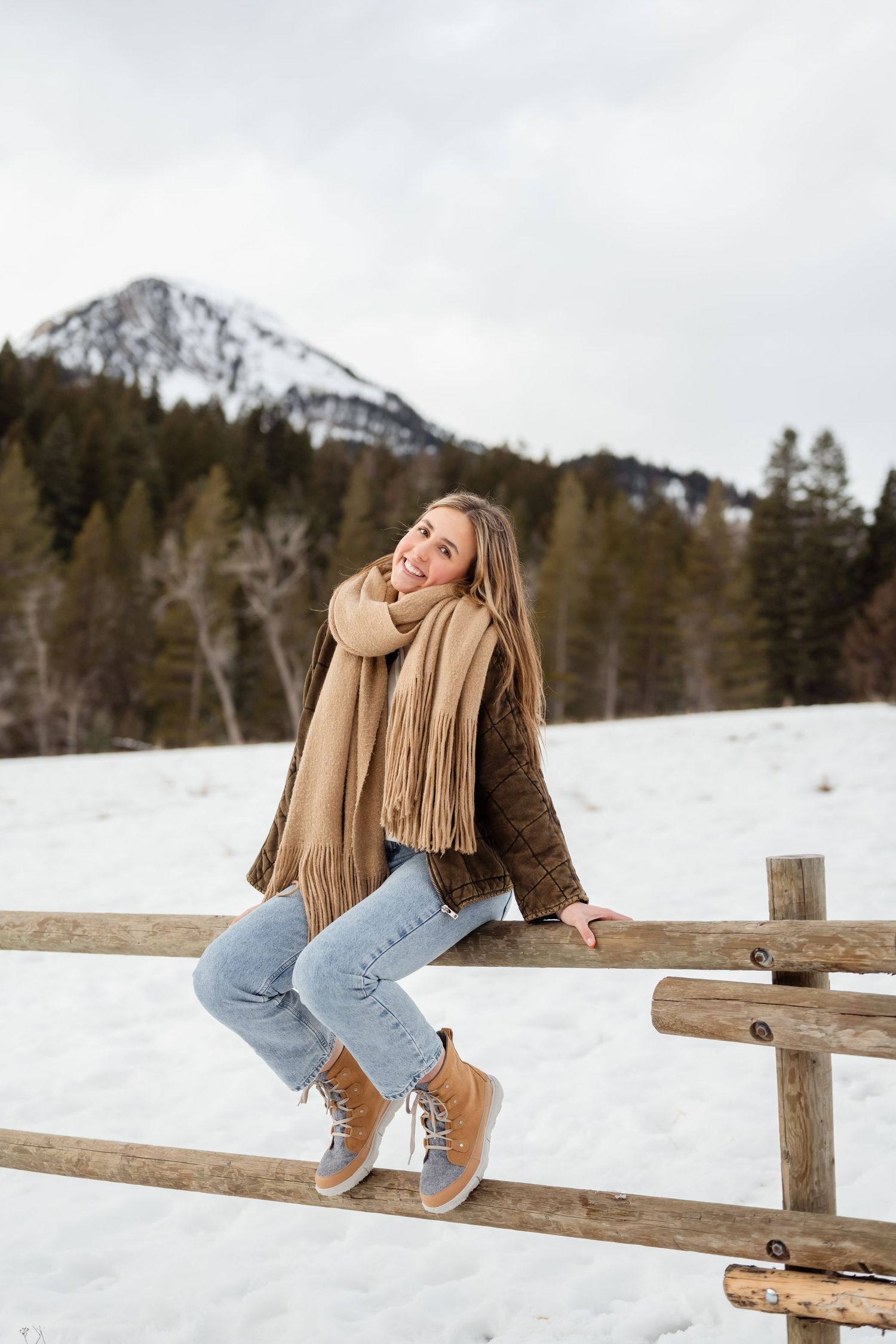 Person in winter attire sits on wooden fence with snowy mountain landscape in background during winter season.