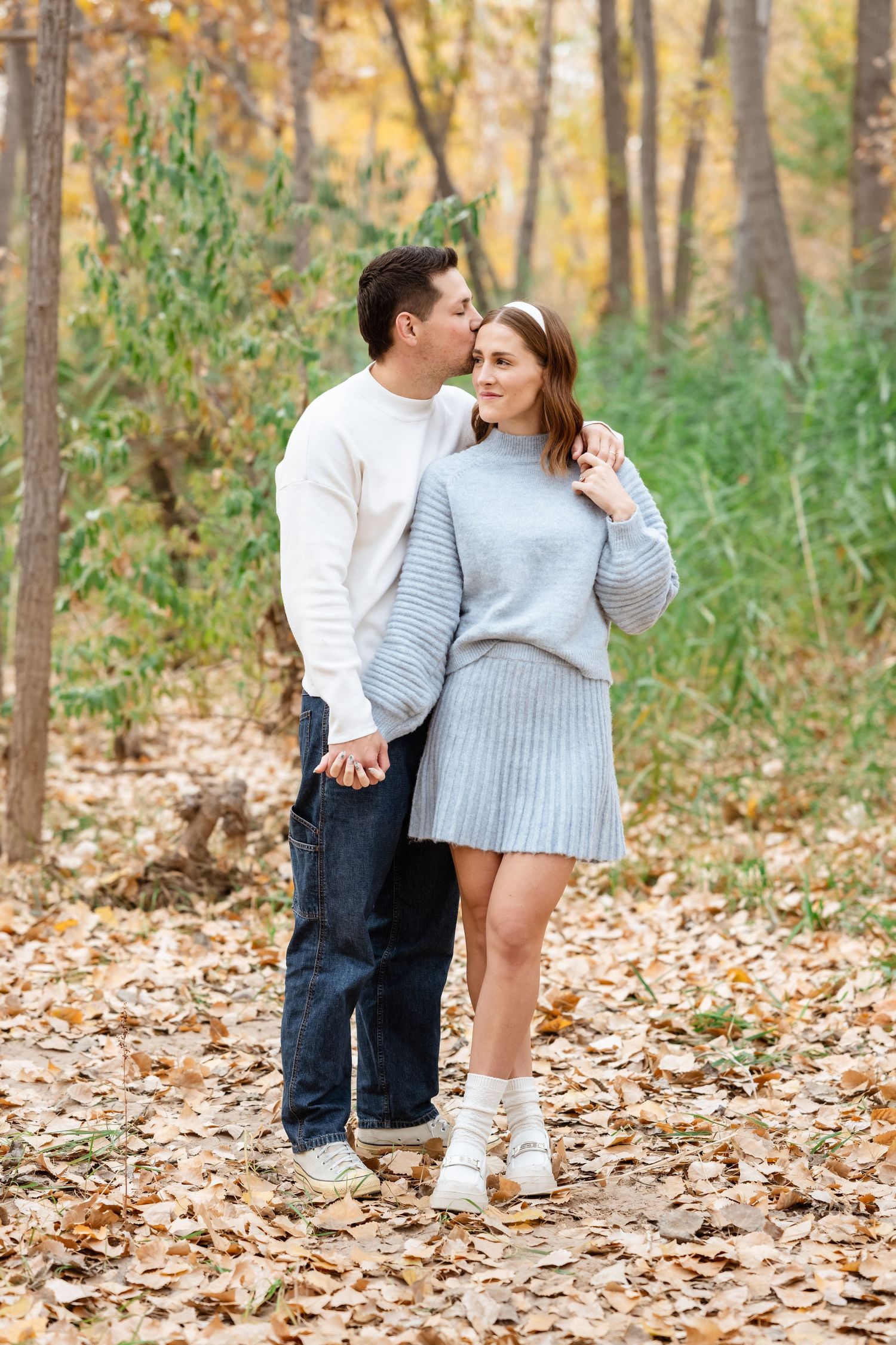 A couple shares an intimate moment while walking through fallen leaves in an autumn forest setting.