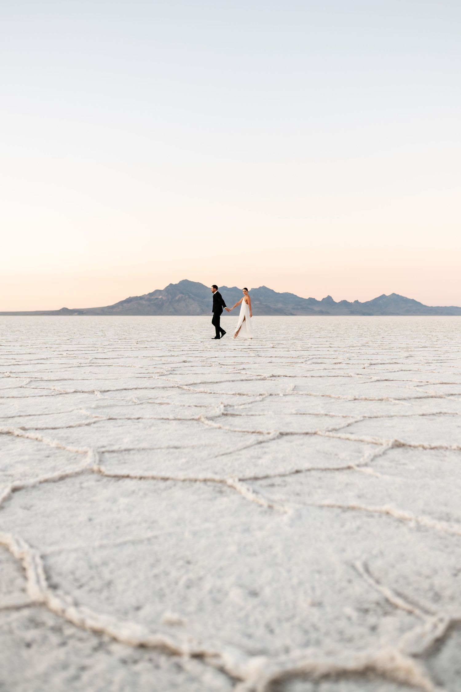 Utah Salt Flats Photographer. Distant silhouette of couple walking on textured salt flats with mountains on horizon.