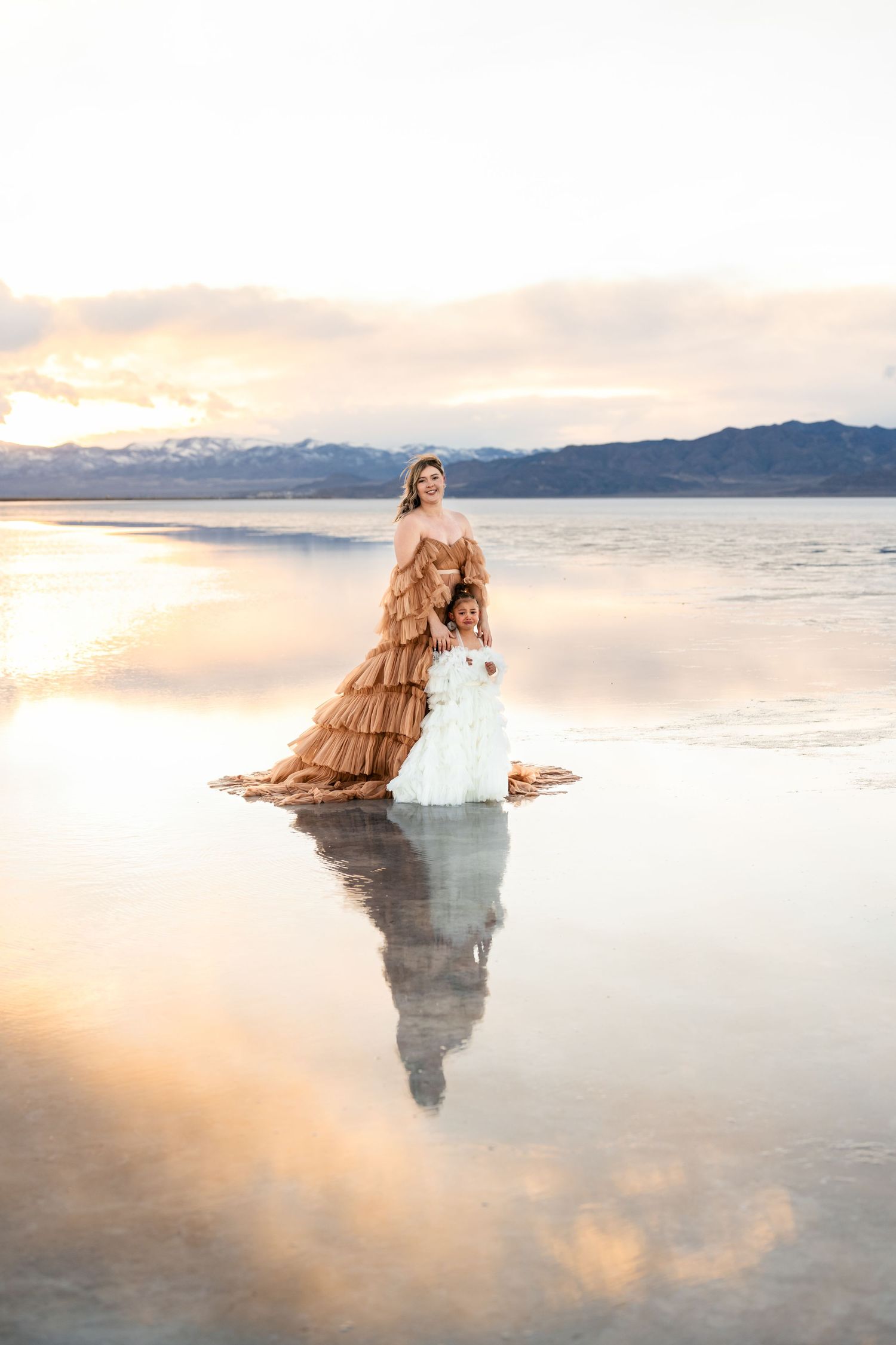 Utah Salt Flats. A figure in a flowing dress stands reflected in the calm waters of a salt flat at sunset with mountains in the distance.