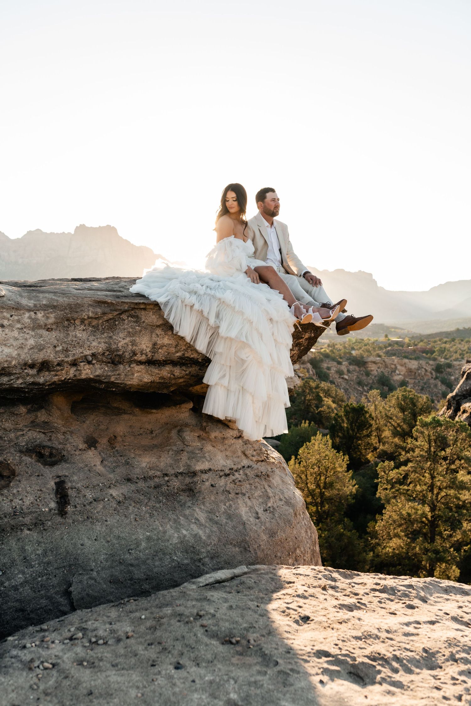 Zion Adventure Session. Couple in wedding attire sits on desert cliff edge at sunset. Utah Zion Photographer.