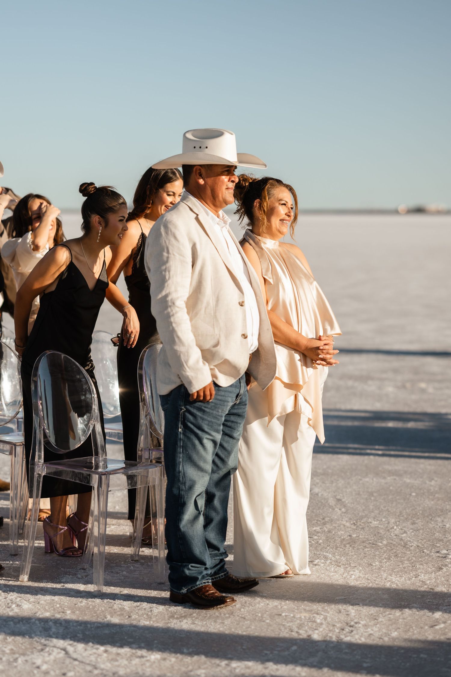 Wedding guests gather on chairs at an outdoor ceremony on a salt flat under bright blue skies.