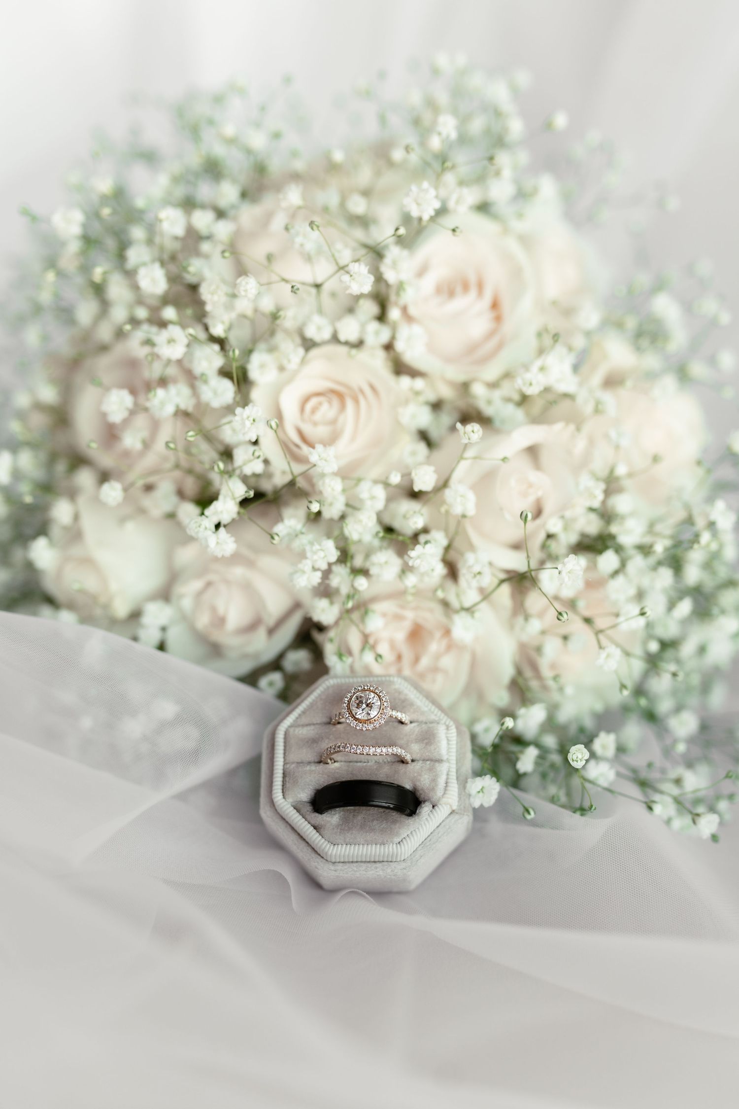 Elegant bridal bouquet of blush roses and baby's breath with wedding rings in clear box on white tulle fabric.
