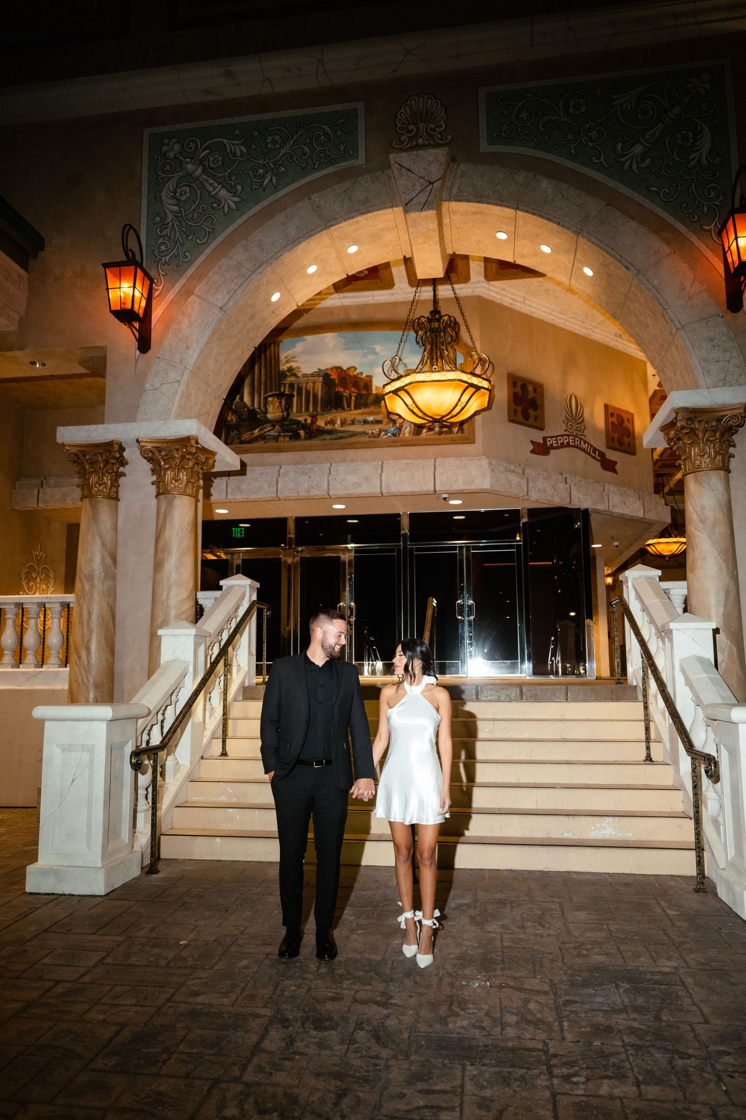 A couple walks through an elegant stone archway corridor with warm lighting and ornate architectural details.