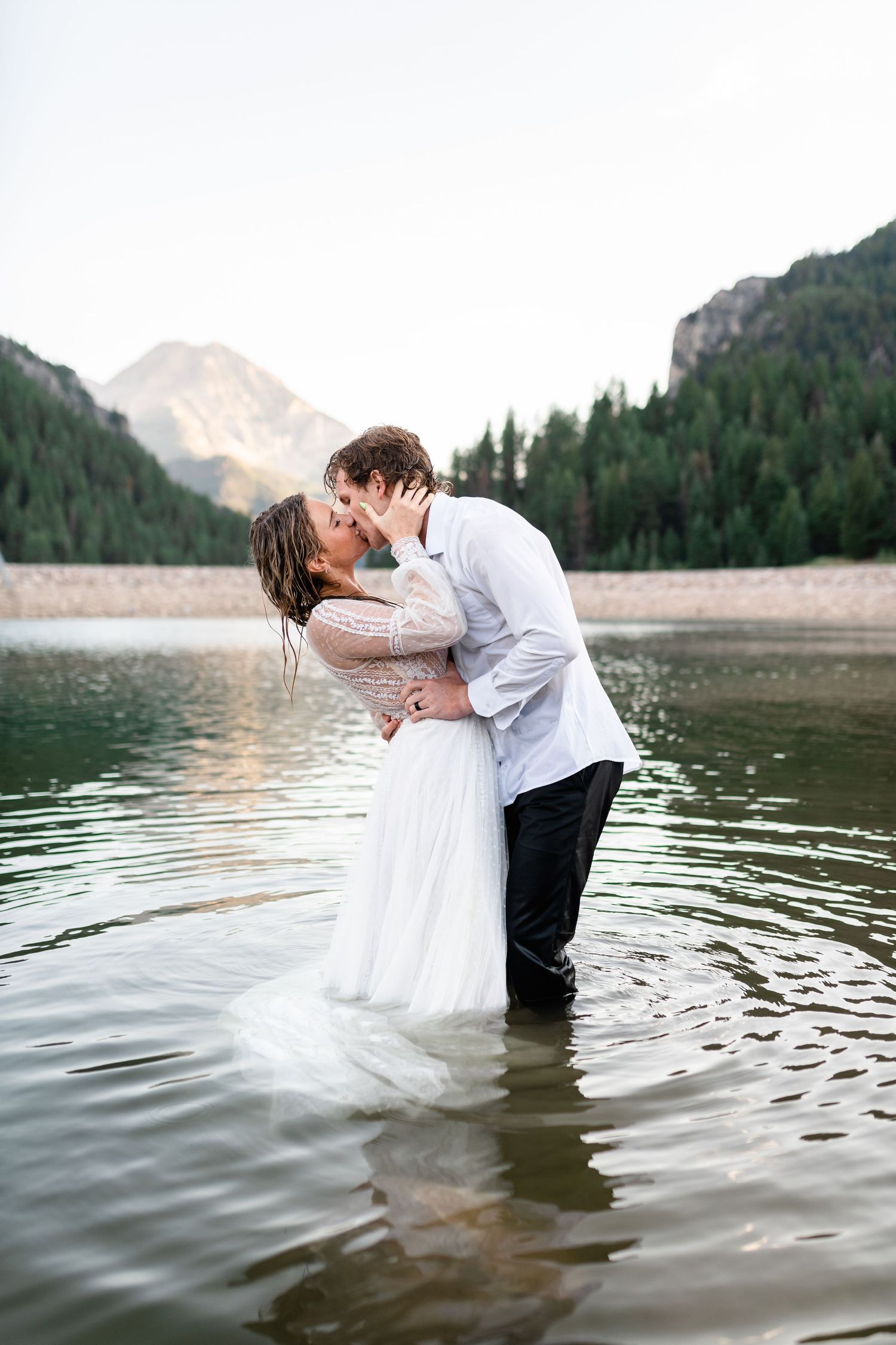 Romantic couple embraces while standing in a mountain lake with evergreen trees in background.  Utah Elopement Photographer.