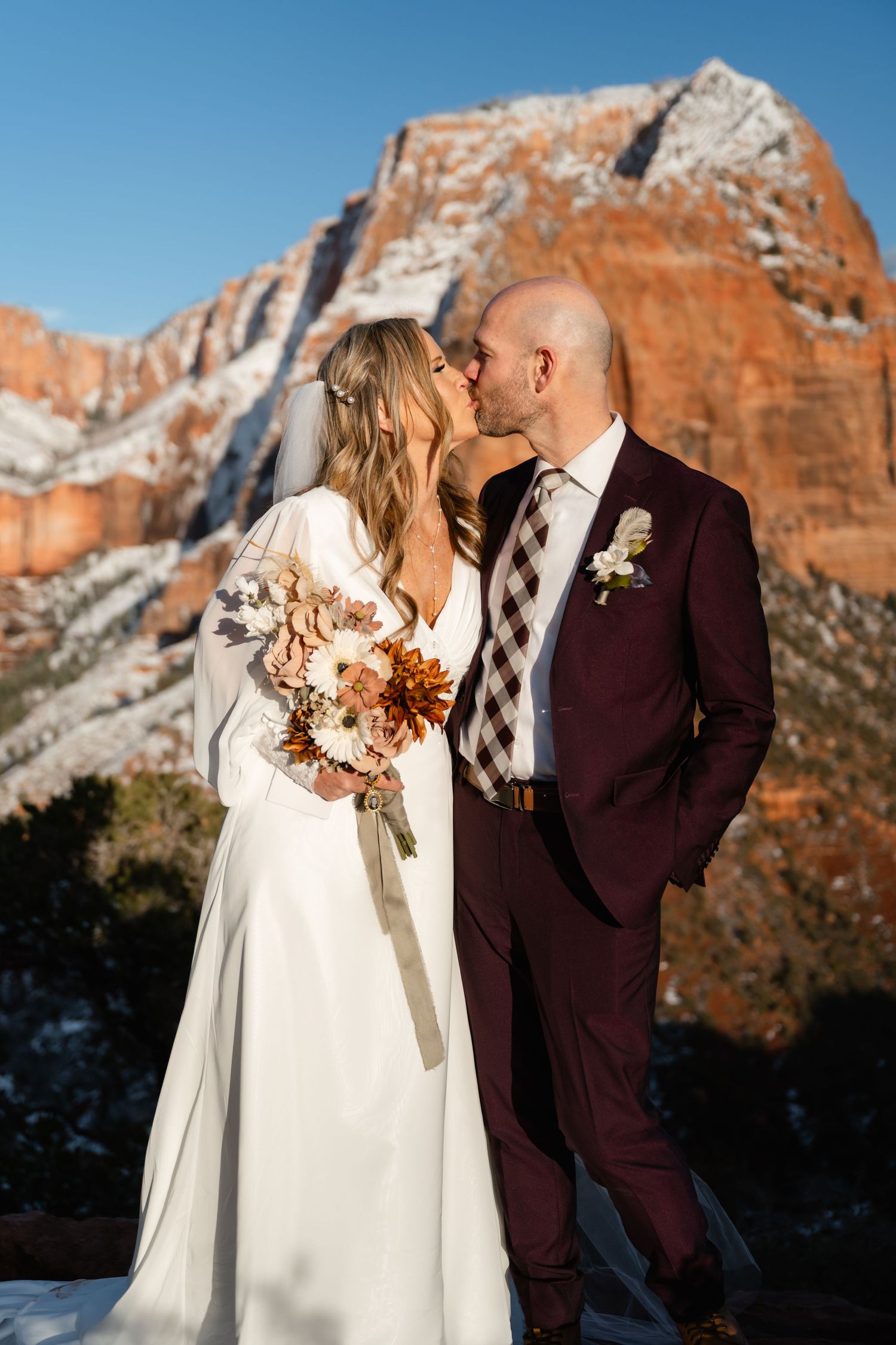 Couple in white dress and burgundy suit standing before majestic red rock cliffs.