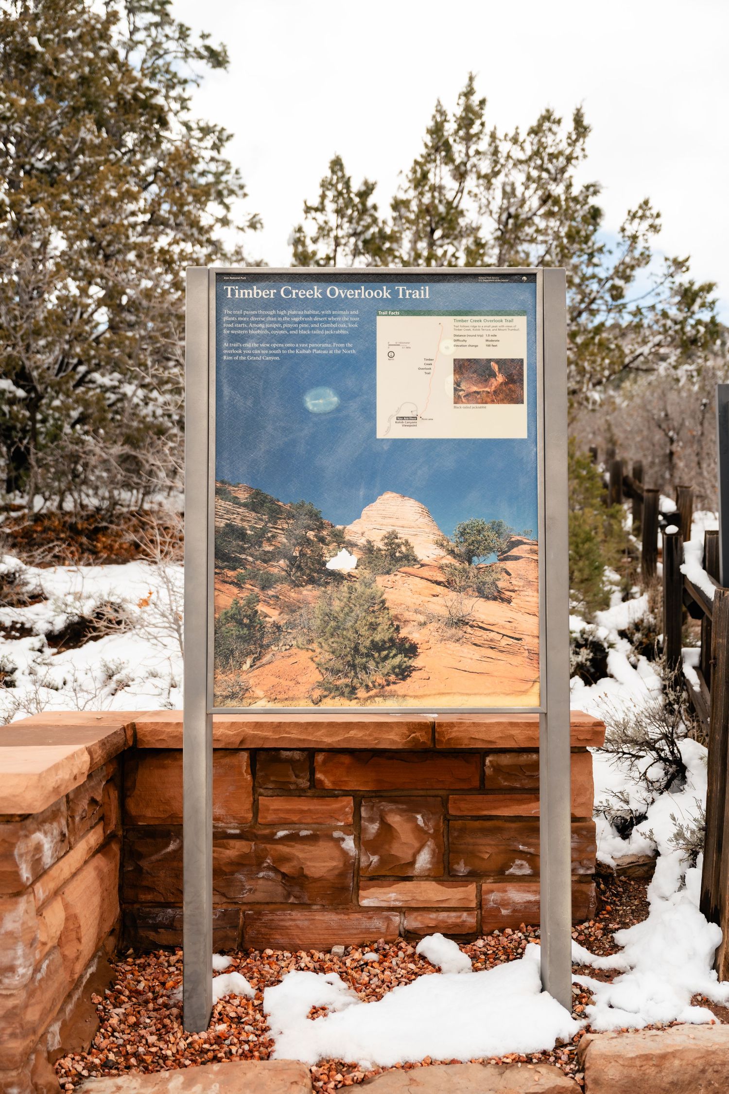 Snow-covered trail sign and information board at Timber Creek Overlook against red rock cliffs in winter.