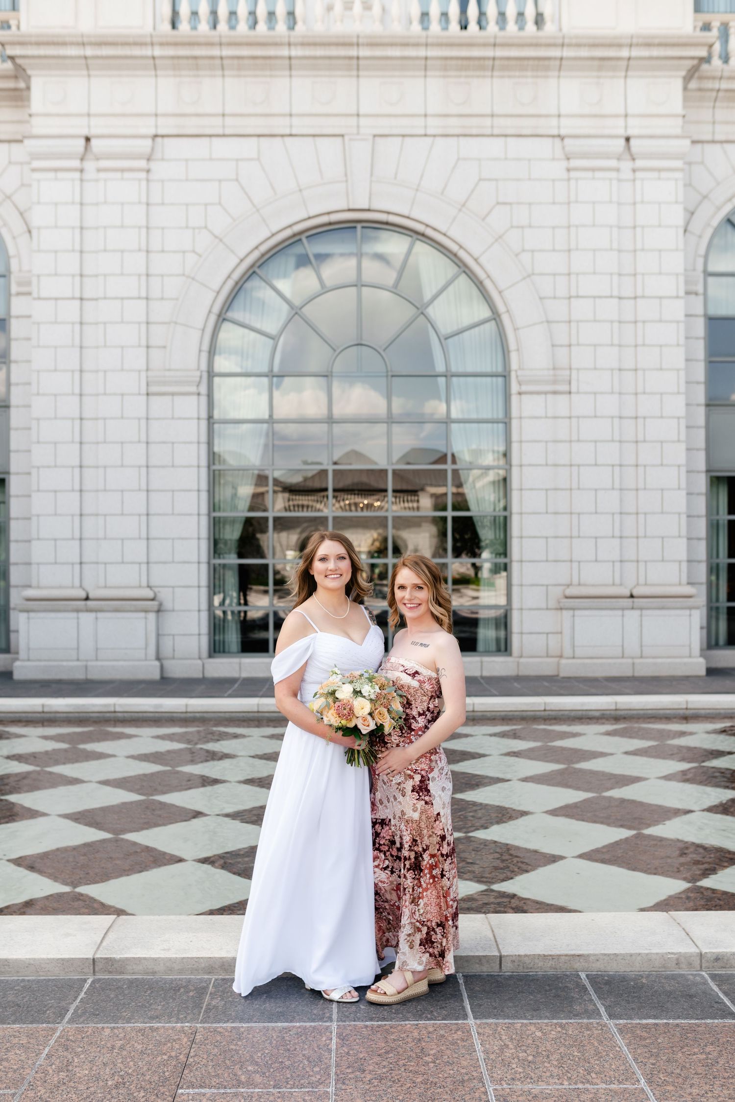 Wedding party poses in front of elegant white stone building with arched windows and checkered marble patio.