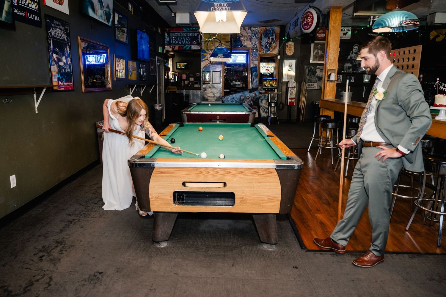 A couple plays pool together in a casual bar setting with neon signs and sports memorabilia on the walls.