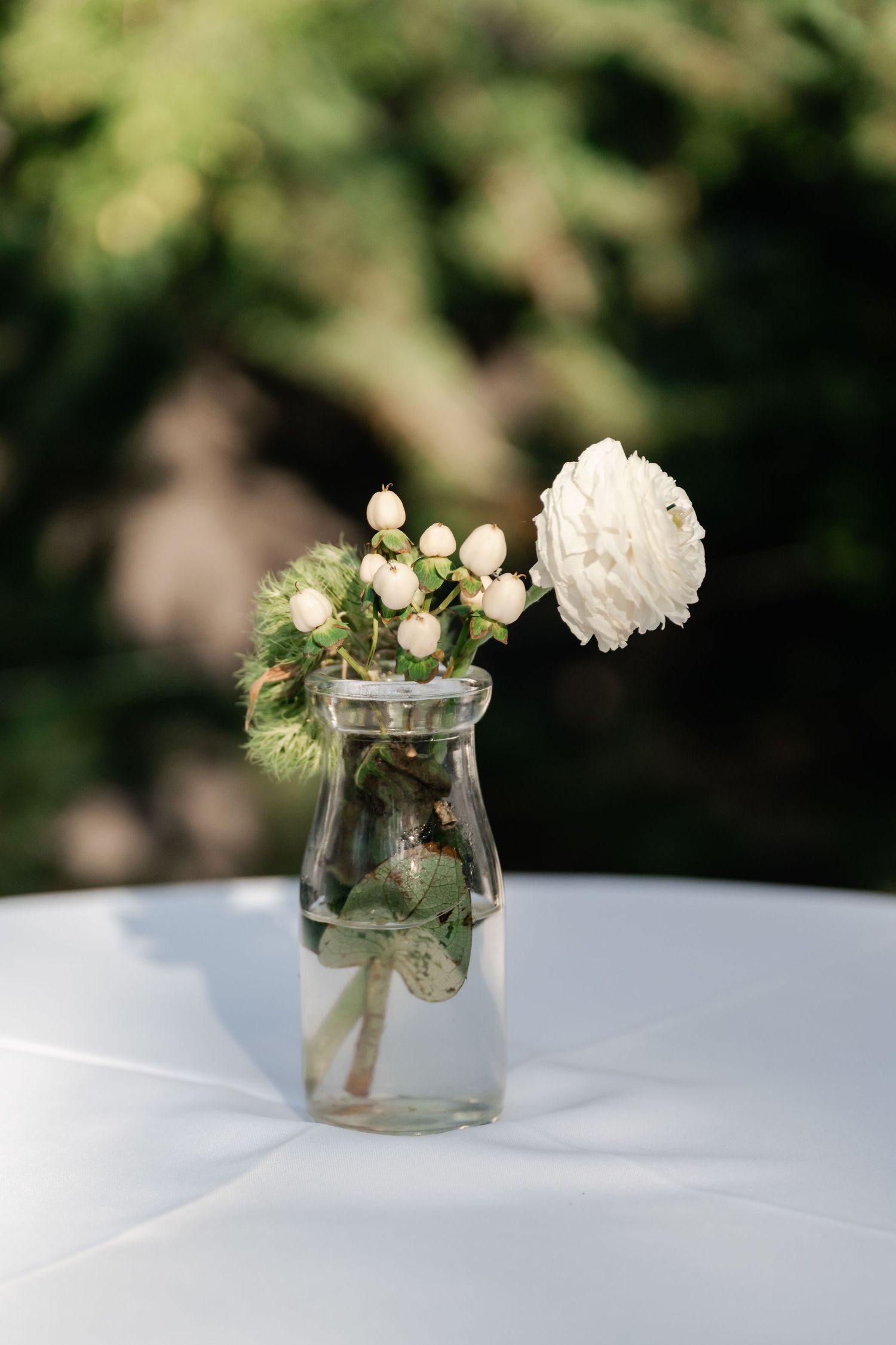 Small glass vases with white flowers and greenery sit on a white tablecloth.
