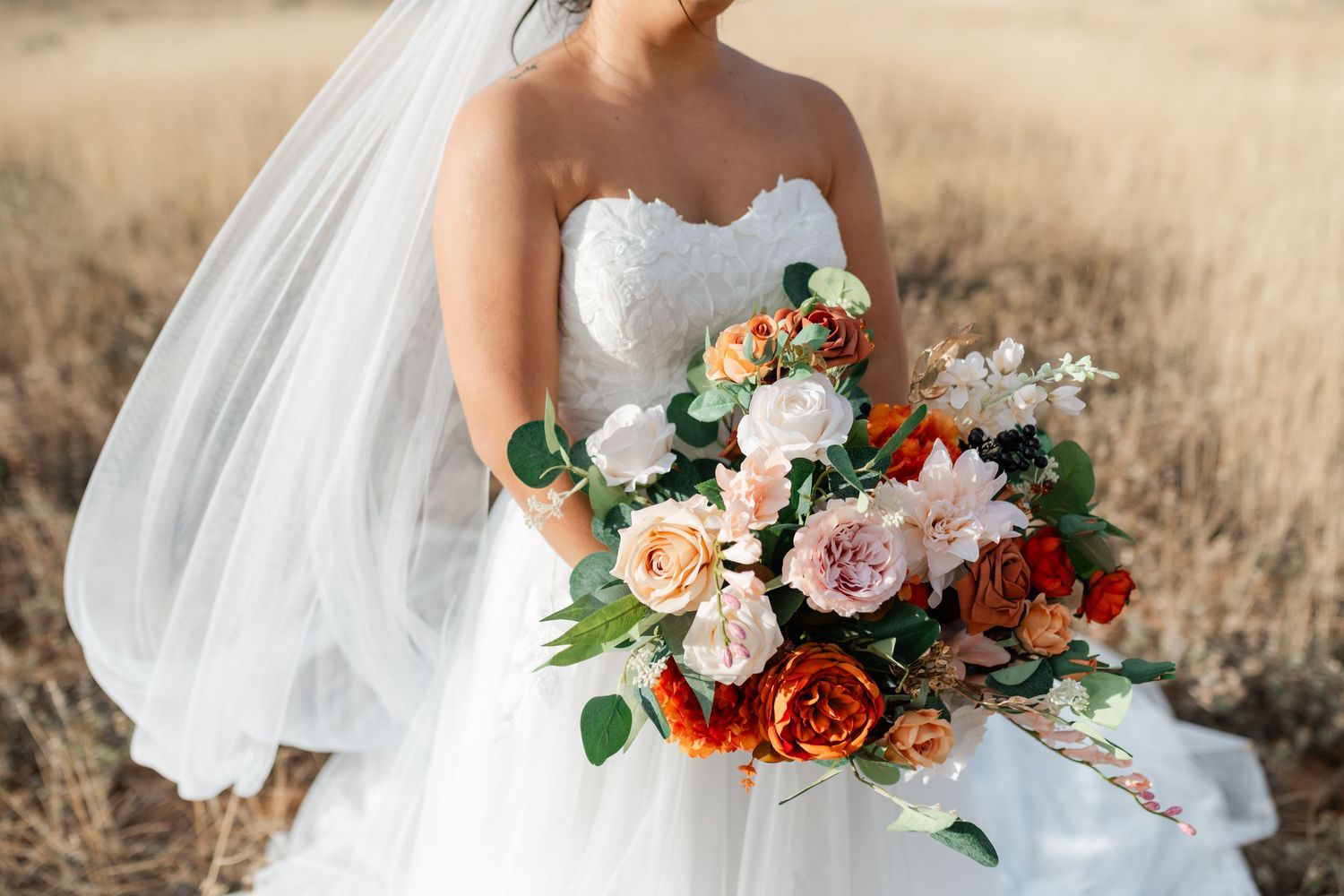Elegant bridal bouquet featuring peach, red, and white roses with flowing white veil in outdoor setting.