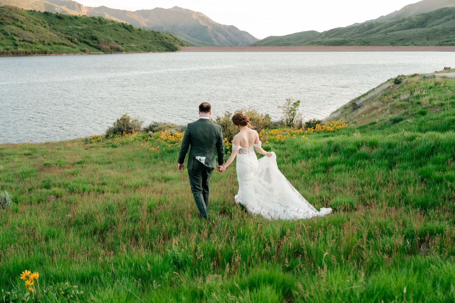 A wedding couple walks through wildflowers with mountains and a lake in the background.