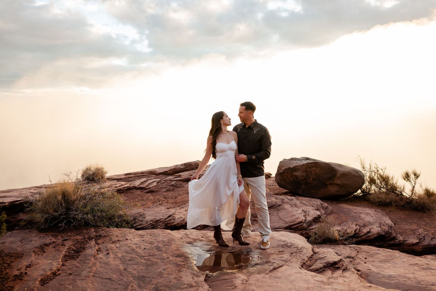 A couple embraces during sunset at a scenic desert overlook with red rock formations in the background.