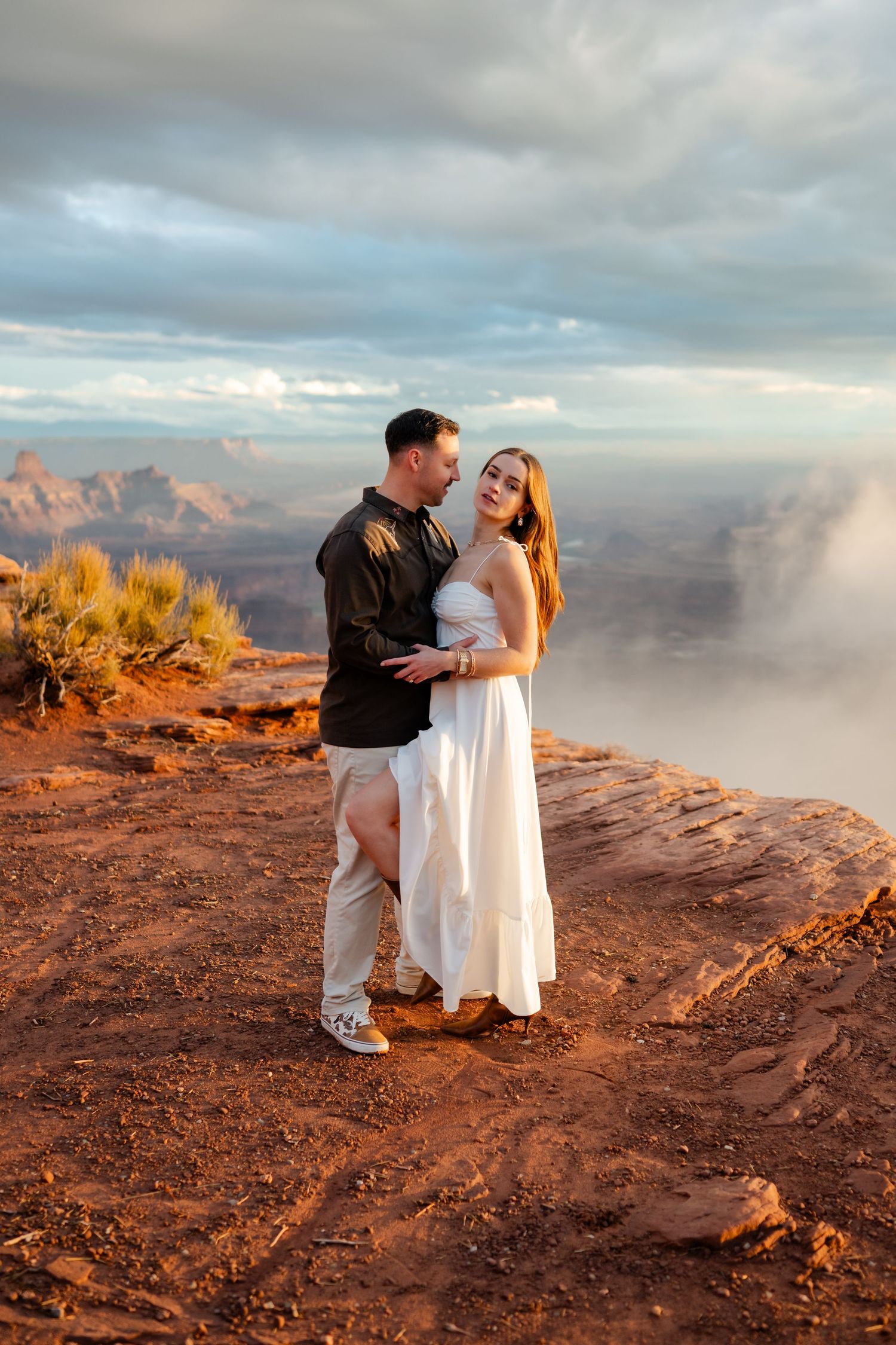 Couple sharing intimate moment on desert cliff edge with golden sunlight and dramatic clouds.