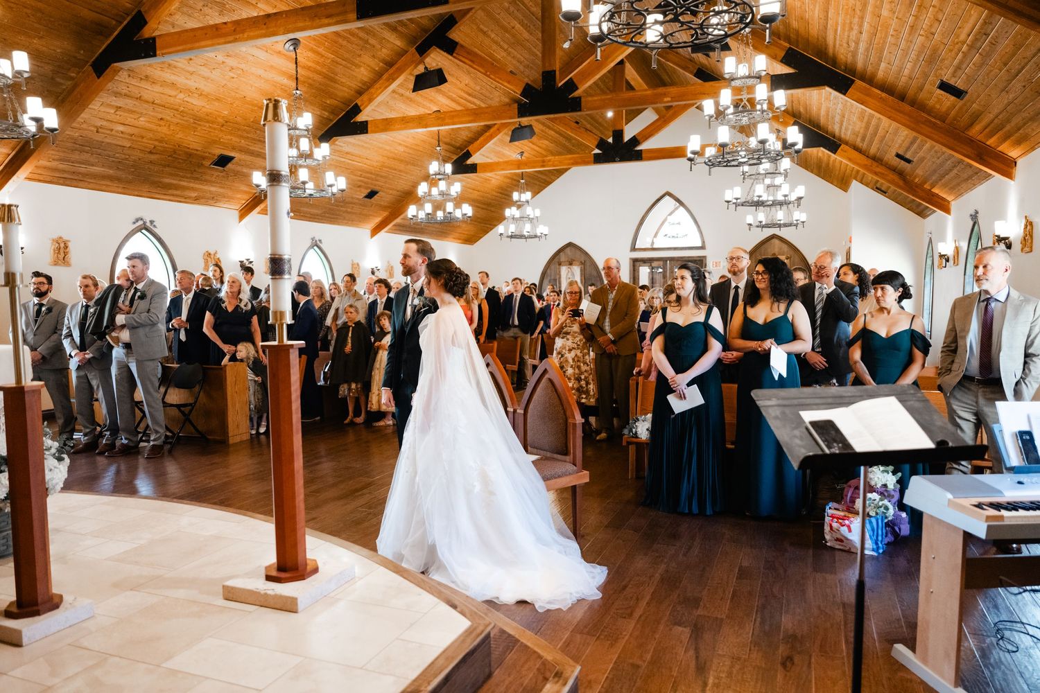 Wedding ceremony in progress inside a chapel with wooden ceiling and chandeliers as guests observe.