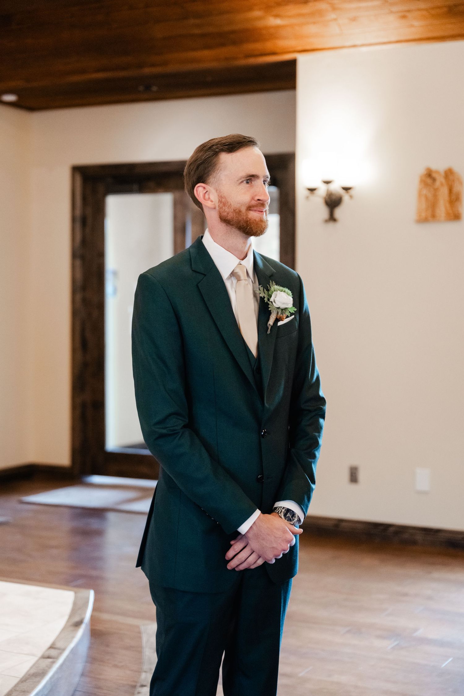 A groom in a dark suit stands in an elegant room with wooden floors and white walls.