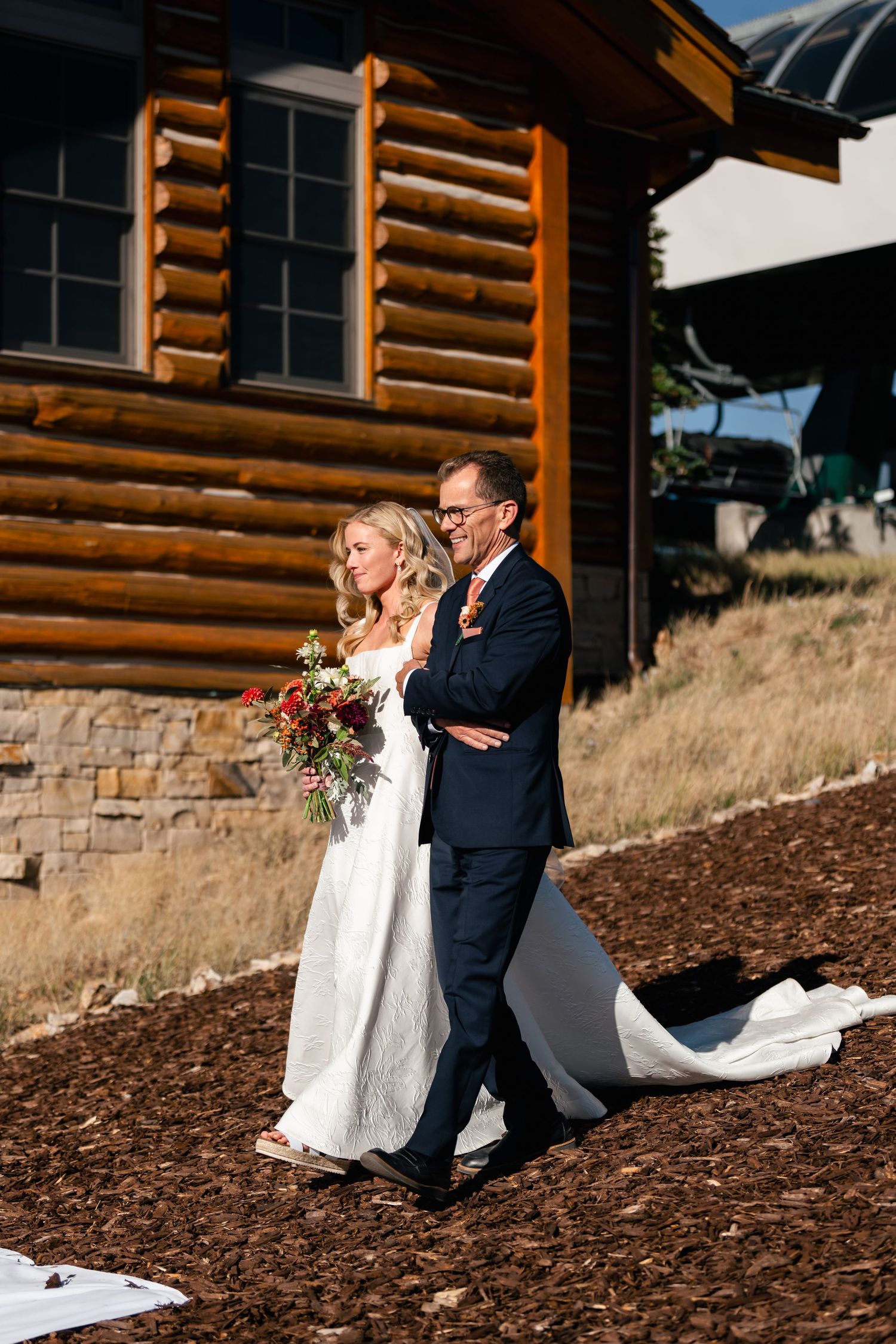 Couple in wedding attire walking along a rustic log cabin exterior with mountain views in the background.