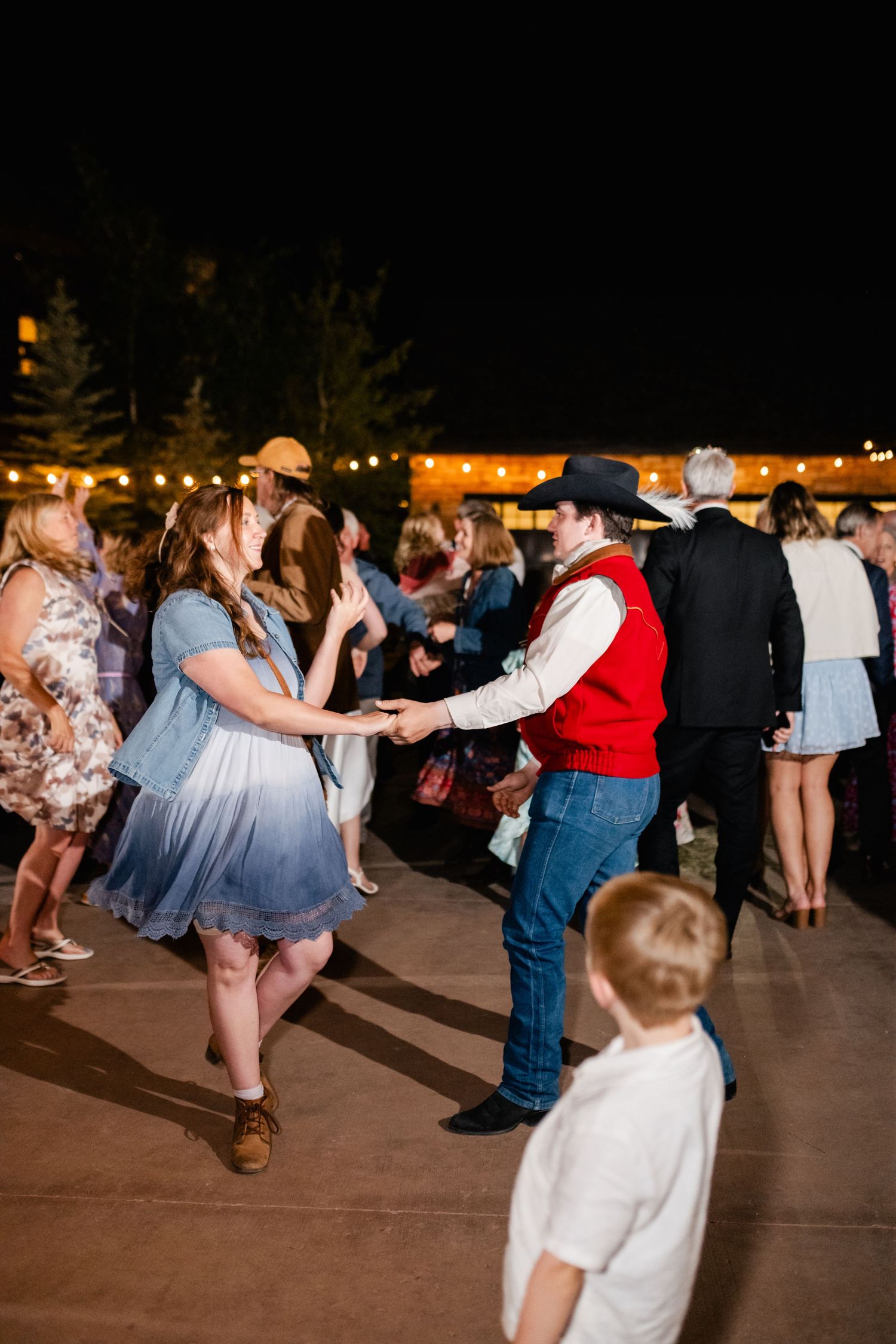 Wedding guests dance together at outdoor reception with string lights overhead.