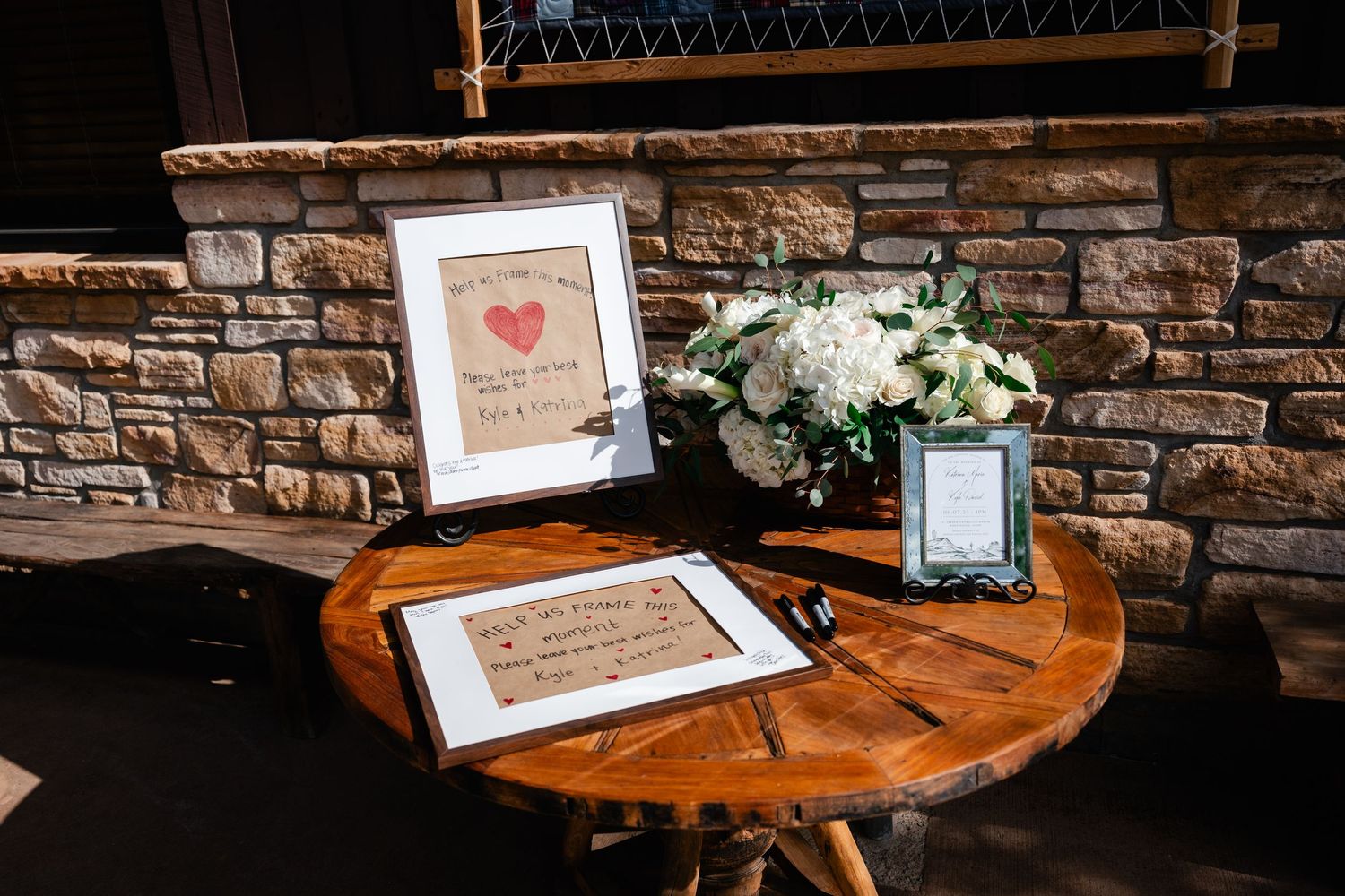 Wedding guest book and decor displayed on rustic wooden table with stone wall backdrop and white floral arrangement.