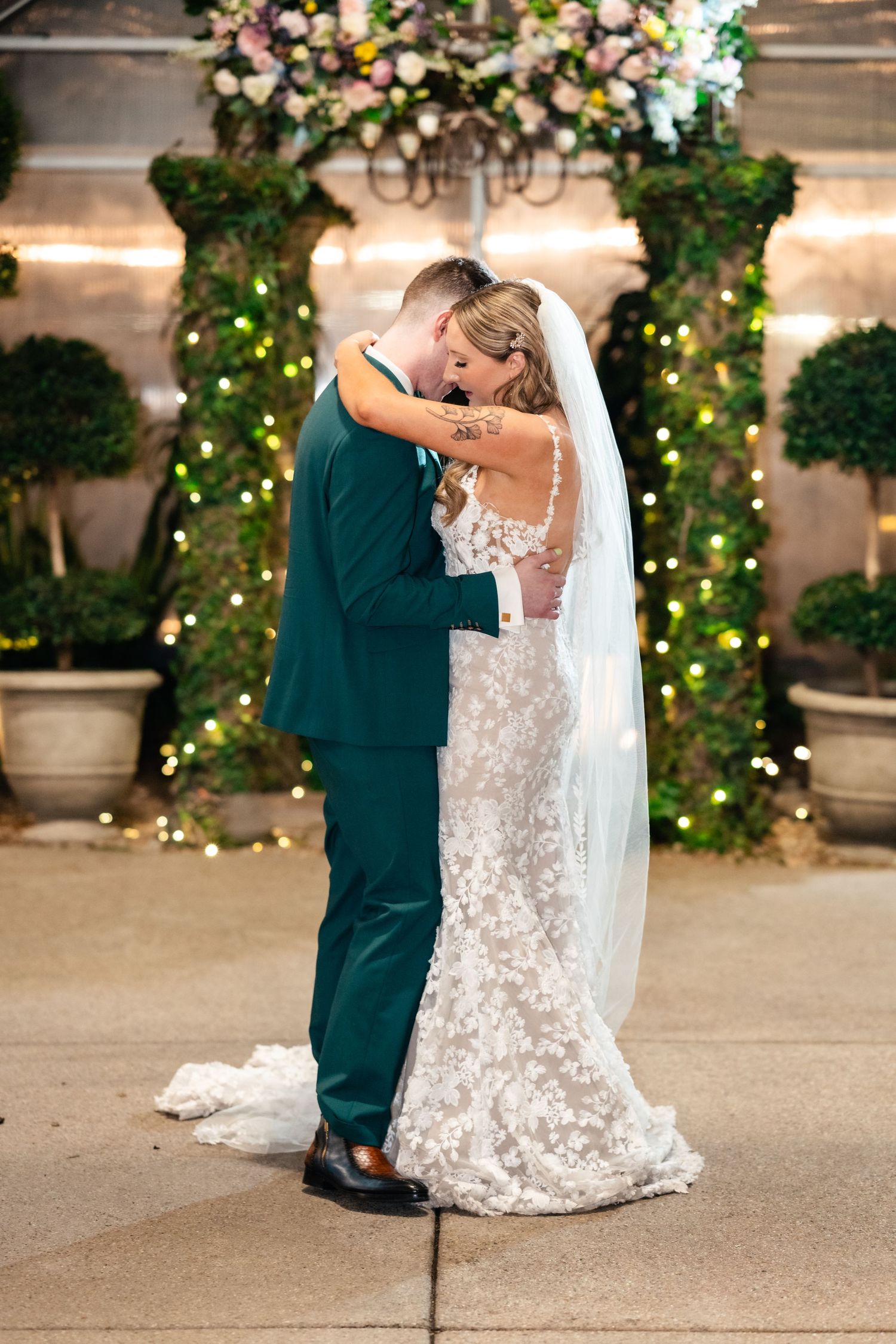 A couple embraces under a floral arch with twinkling lights during their wedding ceremony.