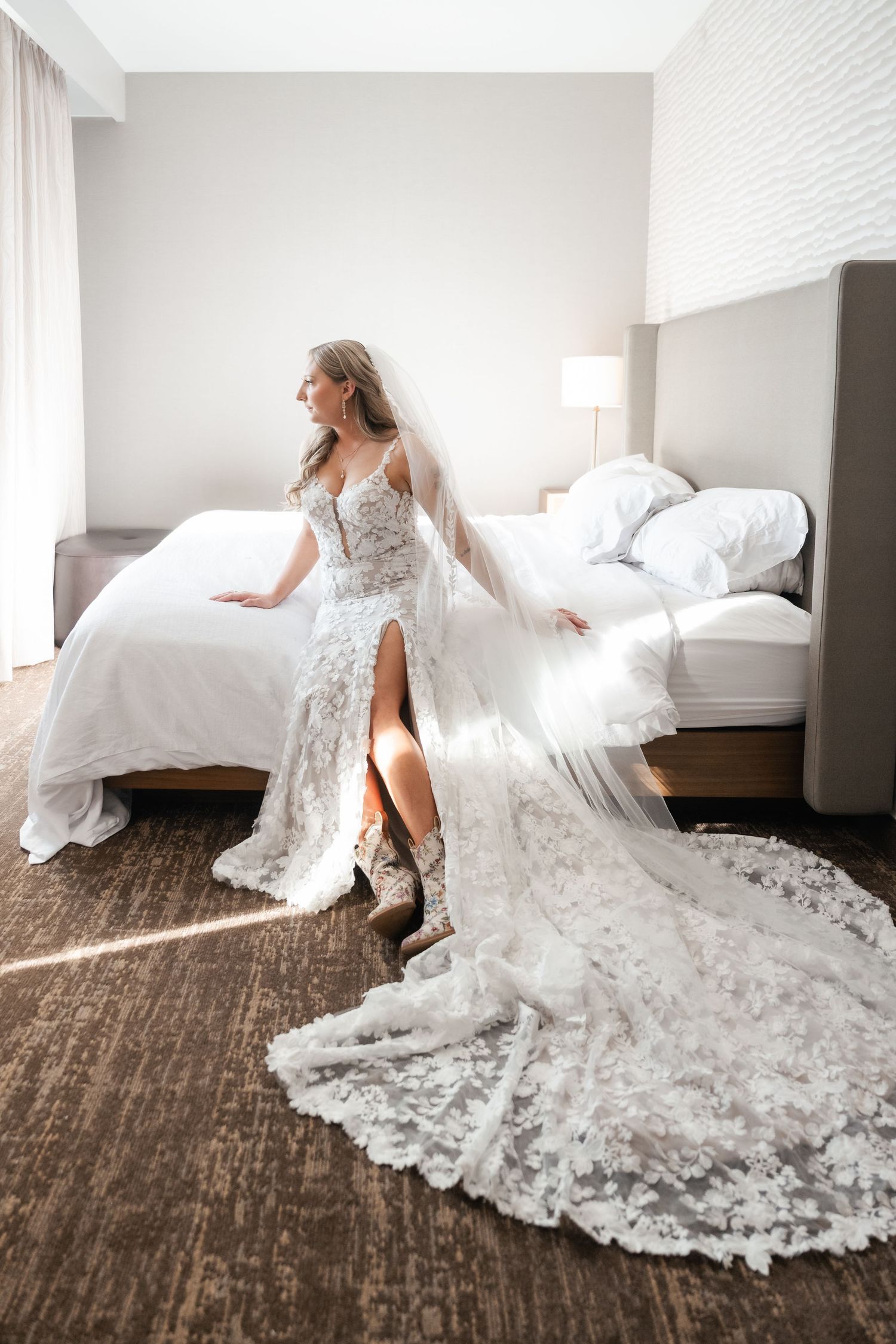 Elegant bride in lace wedding dress with long train poses in modern hotel bedroom.