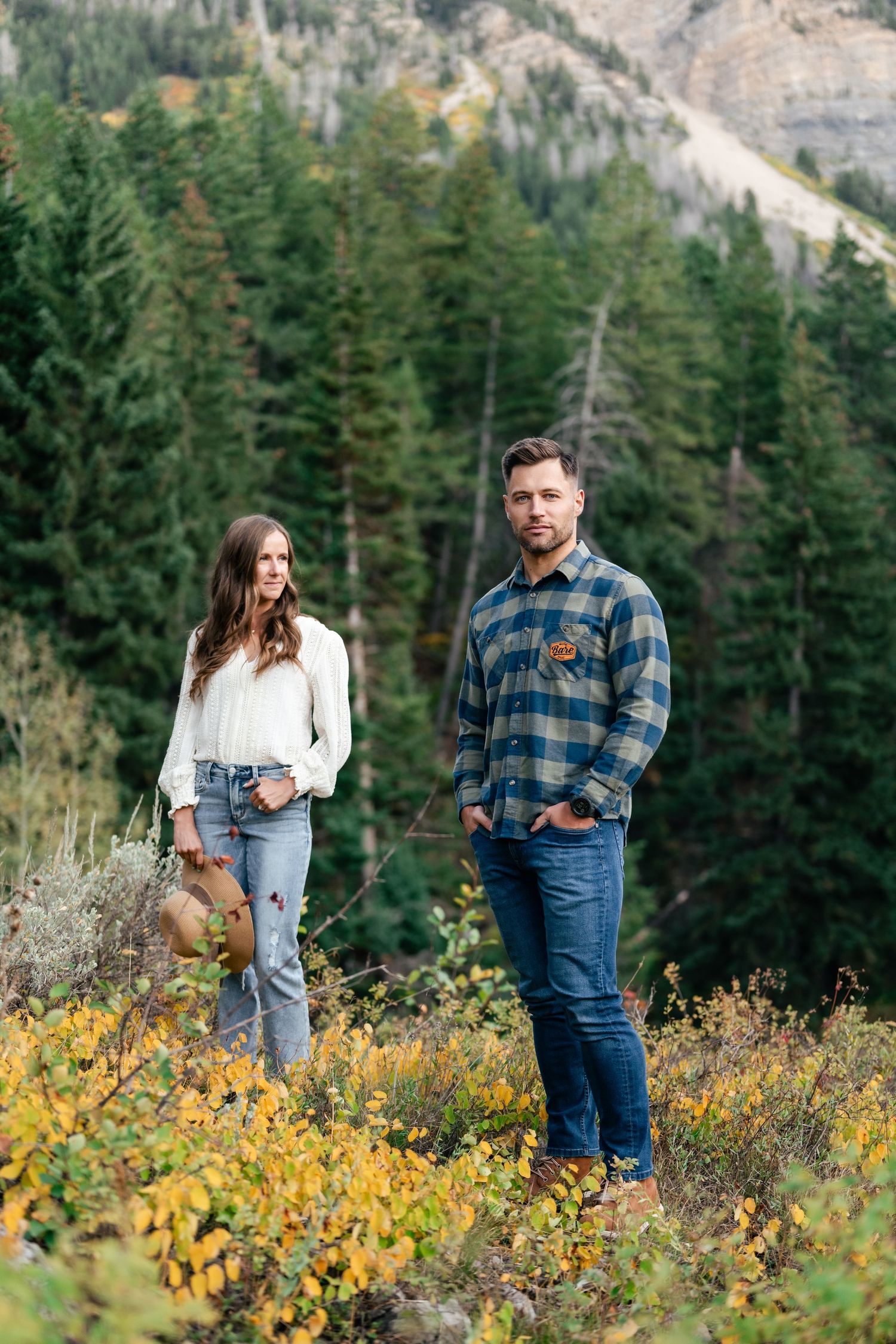 A couple stands among yellow wildflowers with mountain peaks and evergreen forest in the background.