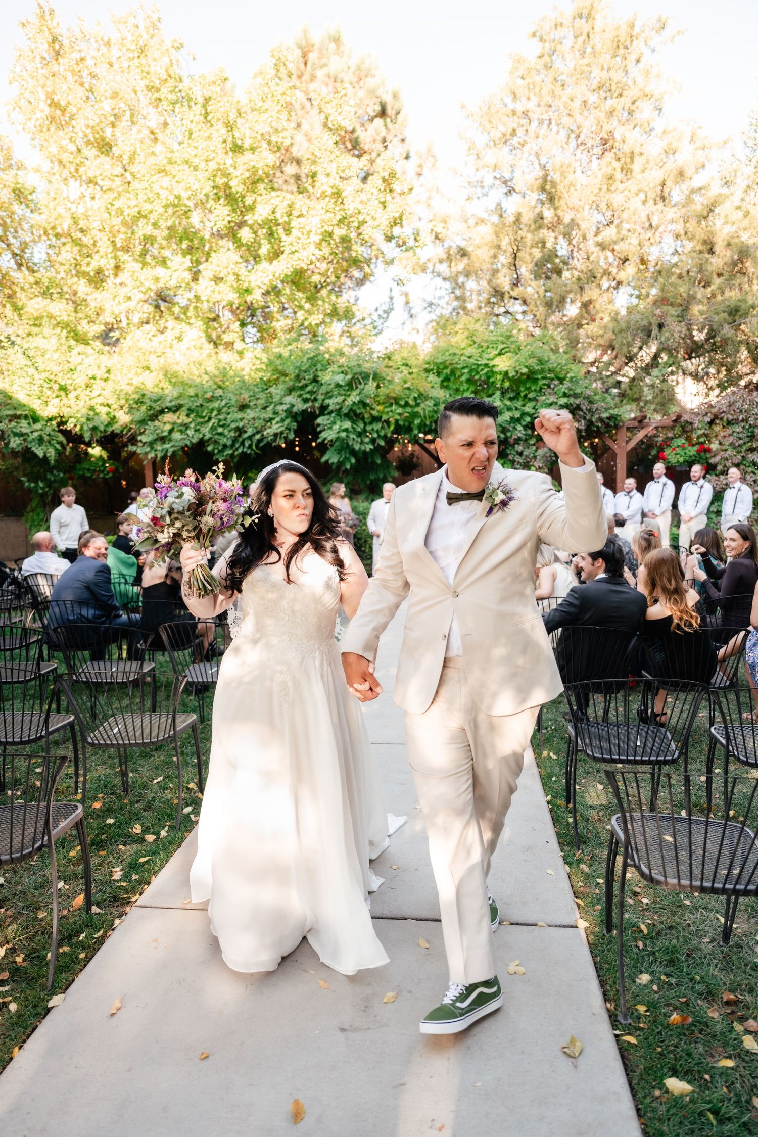 Joyful newlyweds in white attire walk down garden path holding hands after outdoor wedding ceremony.
