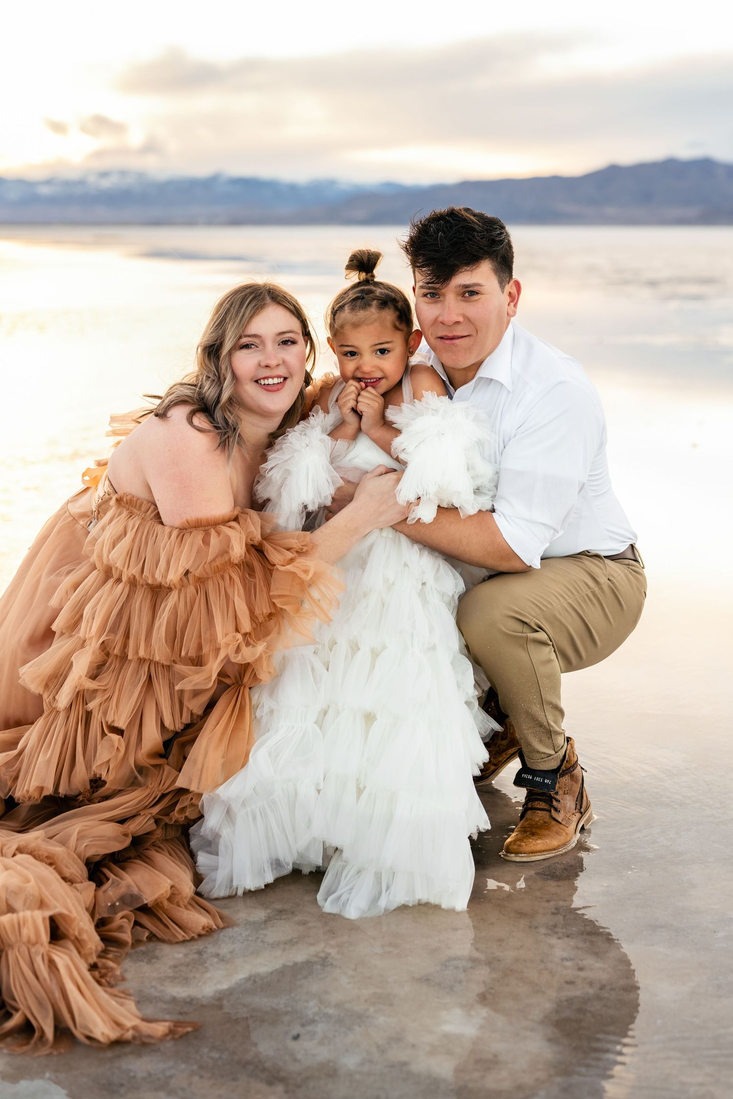 Parents embrace their child affectionately amidst the vast expanse of the flooded salt flats during golden hour.