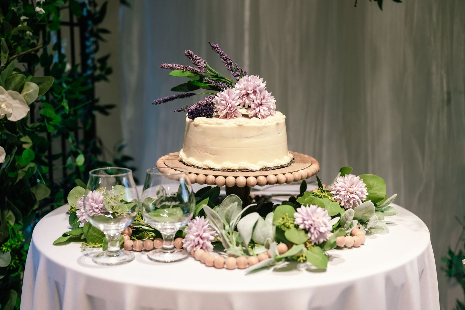 A white wedding cake decorated with pink flowers and greenery sits on a round table with floral arrangements.