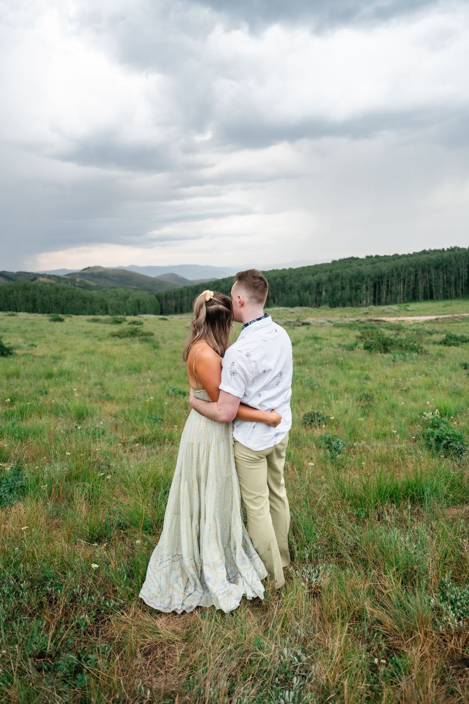 An intimate embrace between newlyweds in an expansive mountain valley with evergreen forest and cloudy skies. Utah mountain engagement.