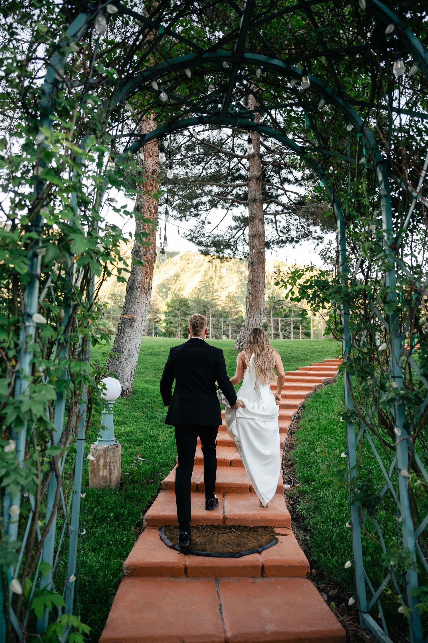 A couple walks through a vine-covered garden archway on terracotta steps leading to an open lawn.