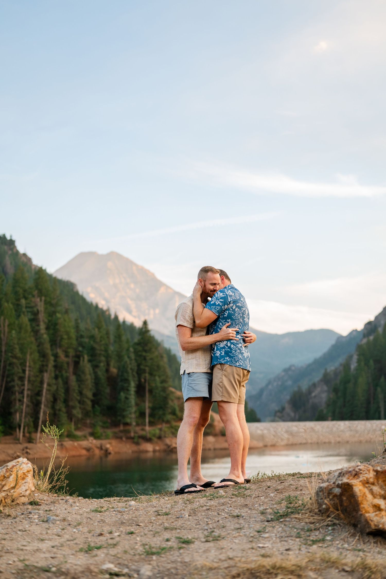Couple embracing by a mountain lake surrounded by pine trees under a clear blue sky.