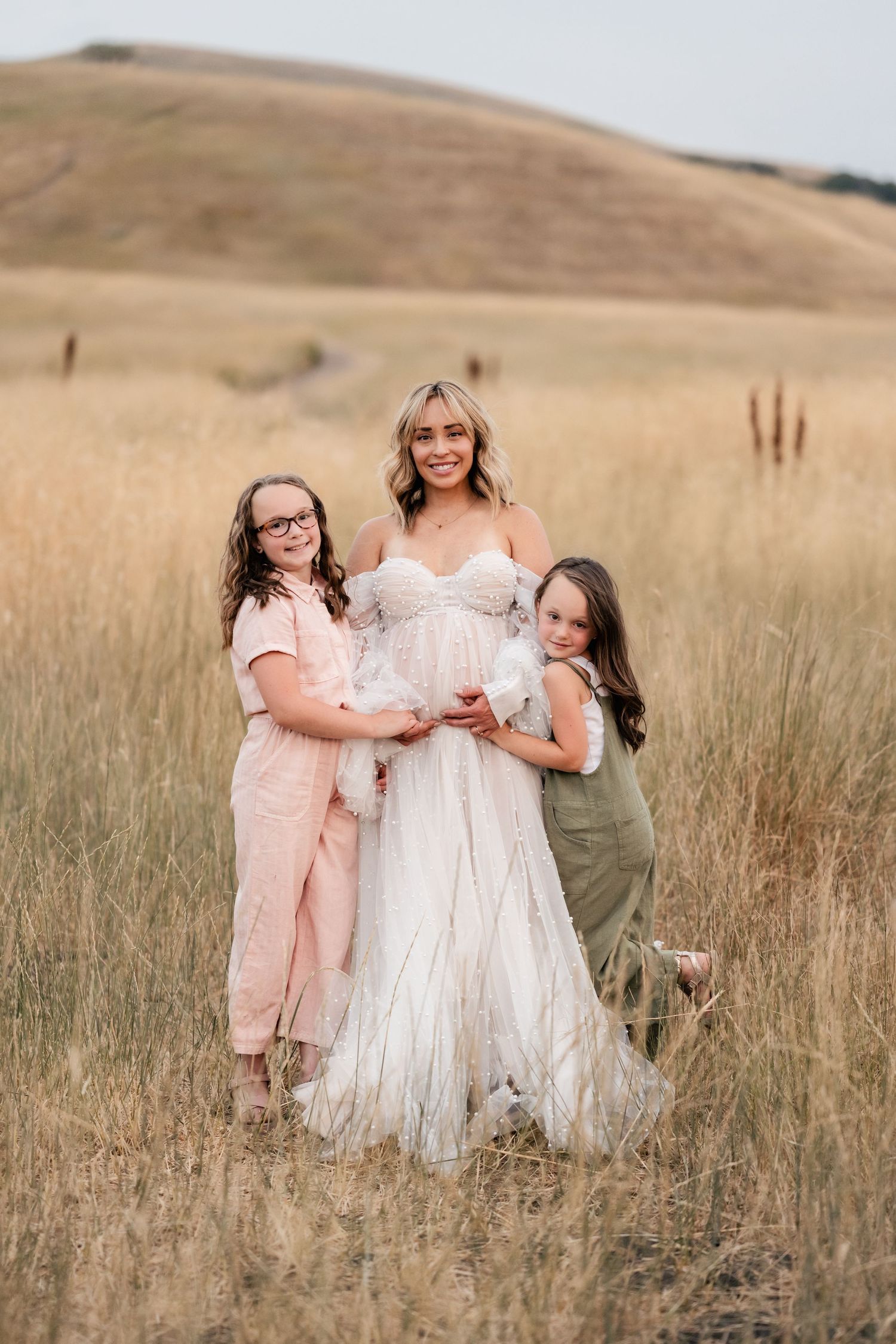 Three people pose together in a sun-drenched field wearing light colored dresses.