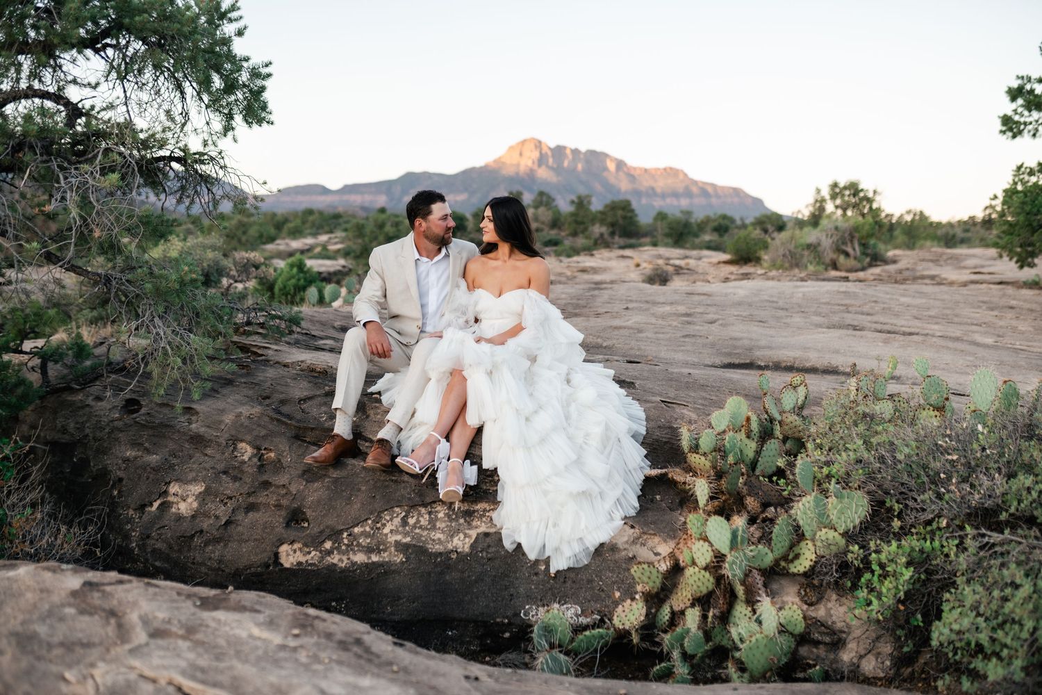 Zion Adventure Session. Romantic couple in wedding attire embraces on desert hillside with mountains and cacti at sunset.