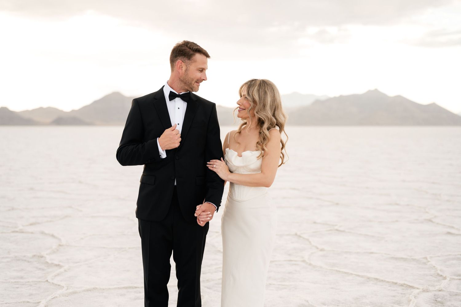 A couple in formal wedding attire dances and embraces on a scenic beach with mountains in the background. Salt Flats Elopement Photographer.