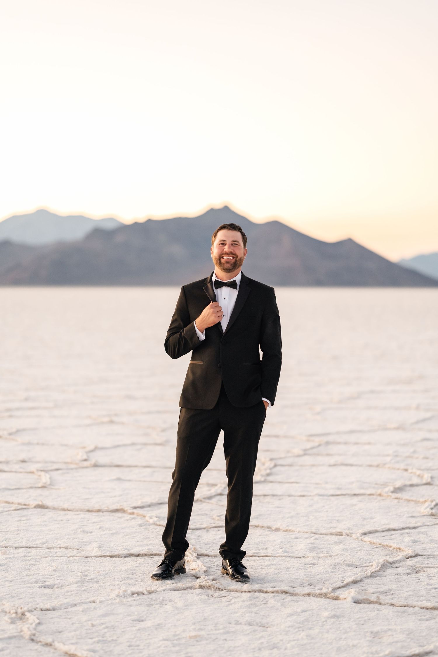 Utah Salt Flats Photographer. Man in black tuxedo poses playfully on white salt flats at sunset with mountains in background.