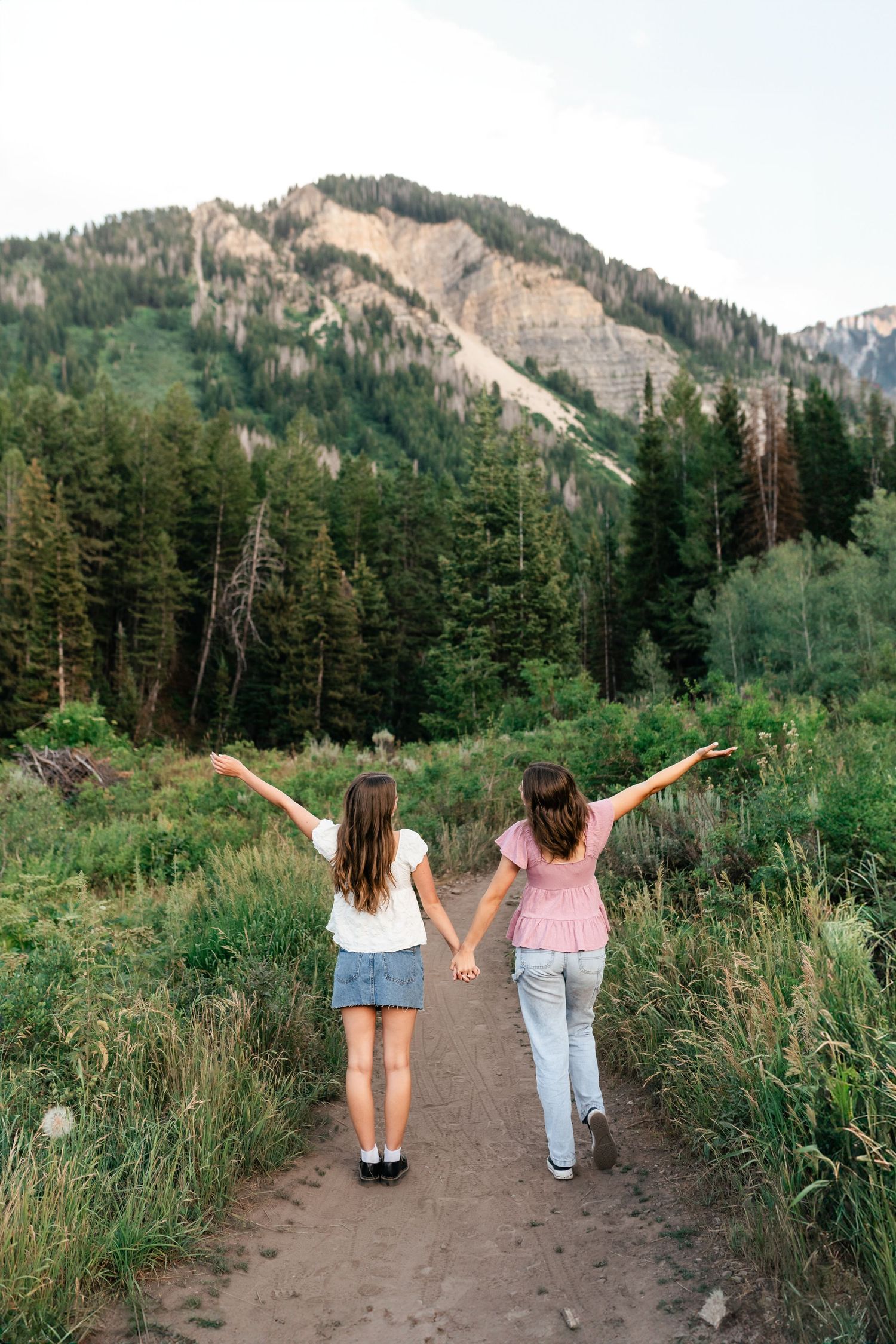 Two friends walking on a mountain trail with arms raised enjoying the scenic mountain landscape and forest views.