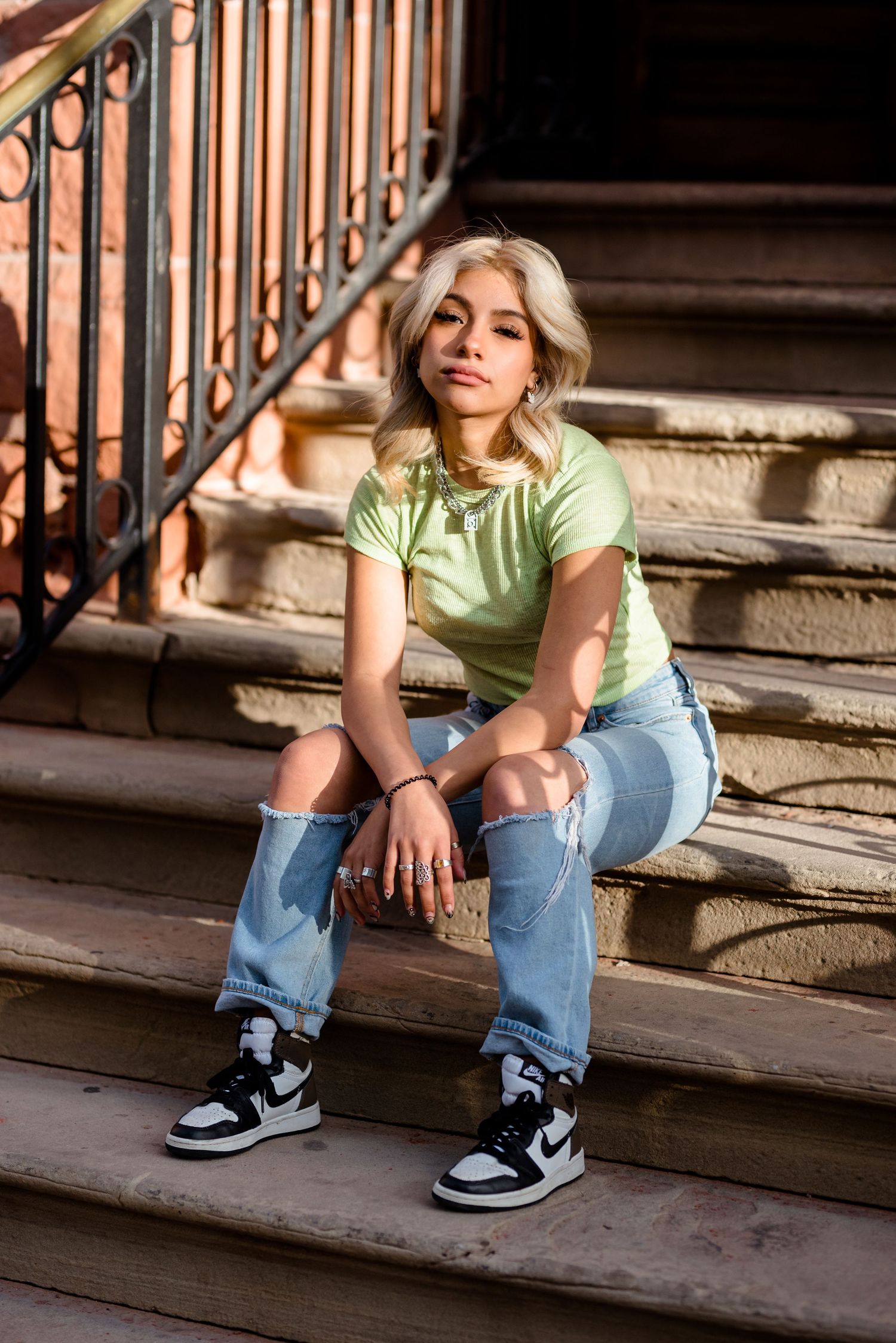 Person sitting on outdoor steps wearing a mint green t-shirt, blue jeans and black sneakers in natural lighting.