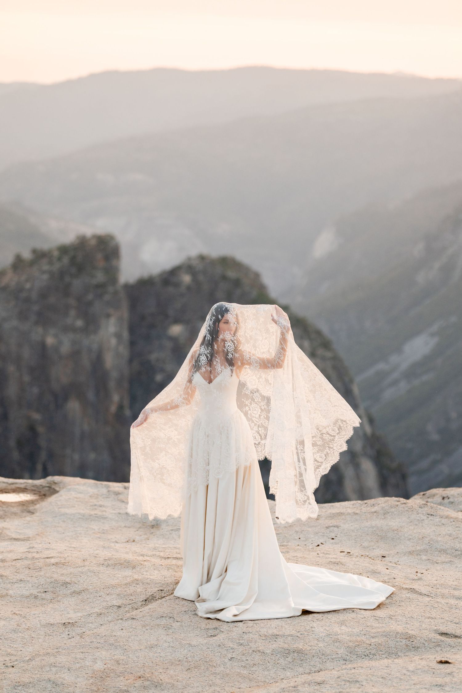 Bride in lace wedding gown with flowing veil lifted by wind on rocky mountain overlook at sunset.